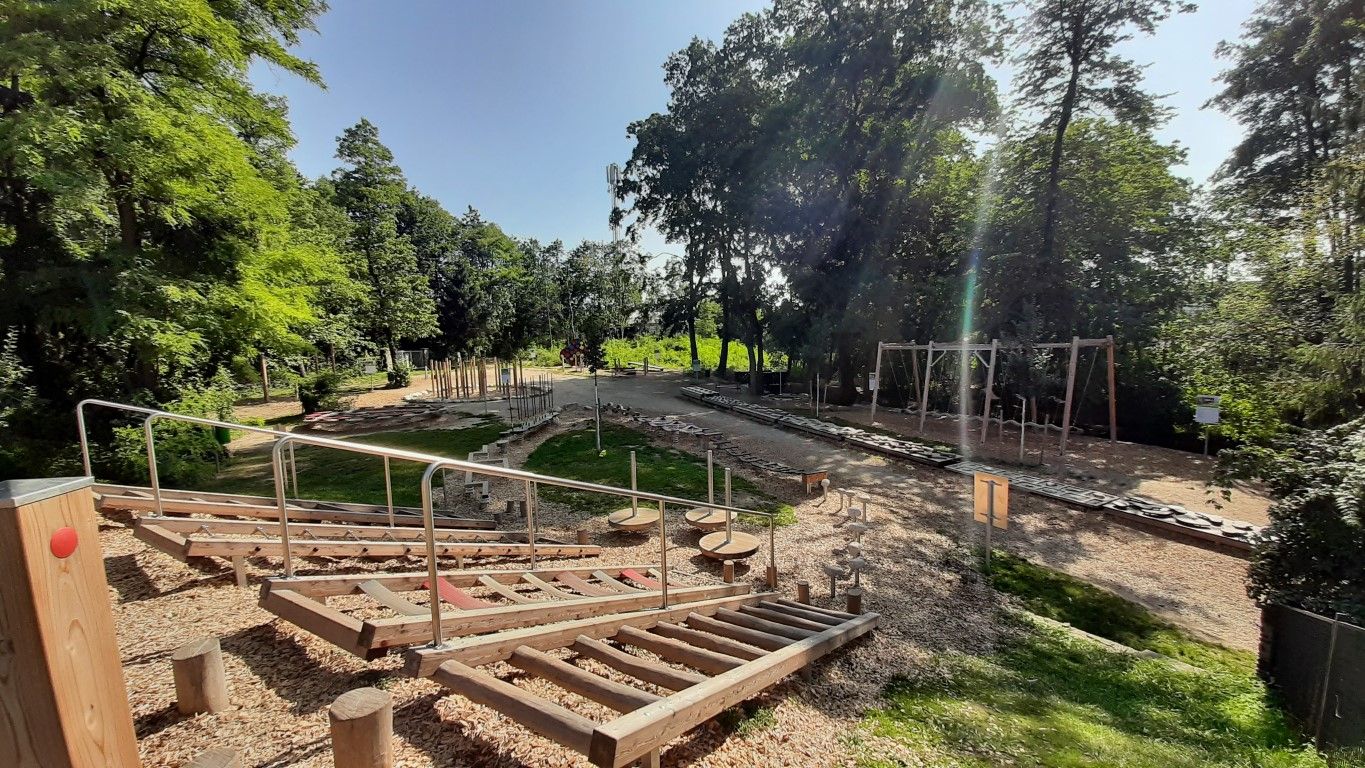 Ein Motorikpark mit verschiedenen Holzelementen und Klettergeräten, umgeben von Bäumen und Grünflächen.