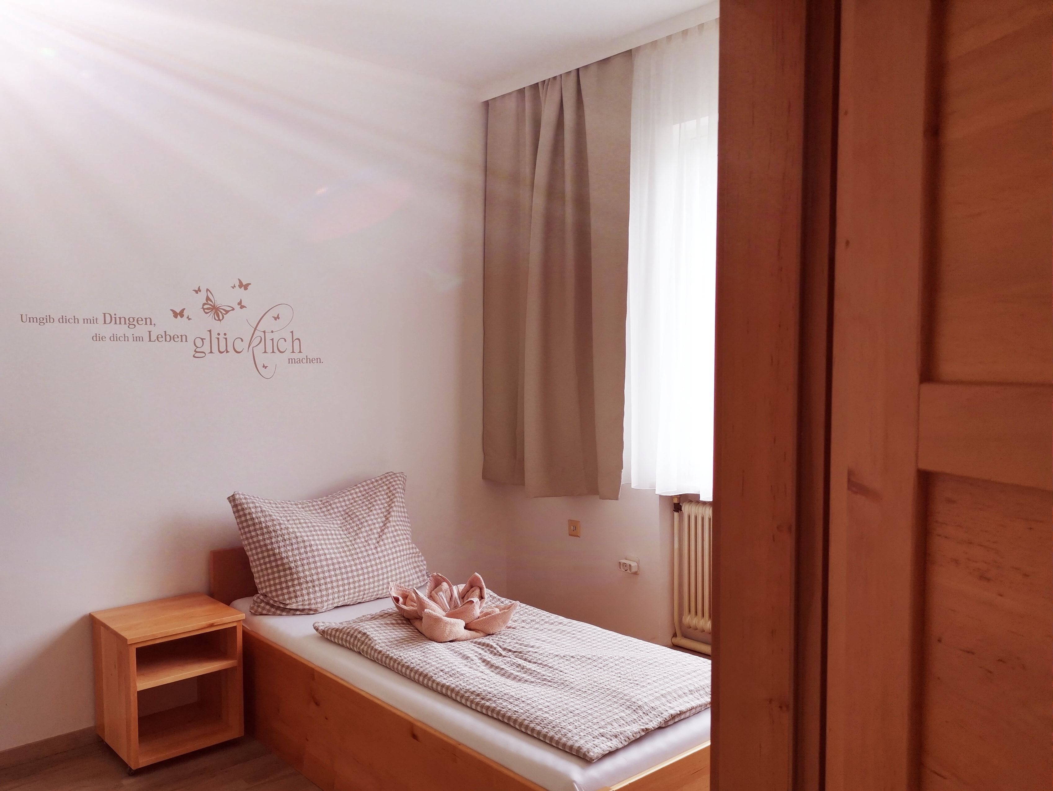 Ein einfaches Gästezimmer mit einem Einzelbett, Holzregal und einem Zitat an der Wand.