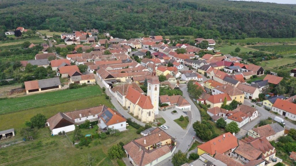 Luftaufnahme eines kleinen Dorfes mit einer zentralen Kirche und roten Dächern, umgeben von grünen Feldern und Wäldern.