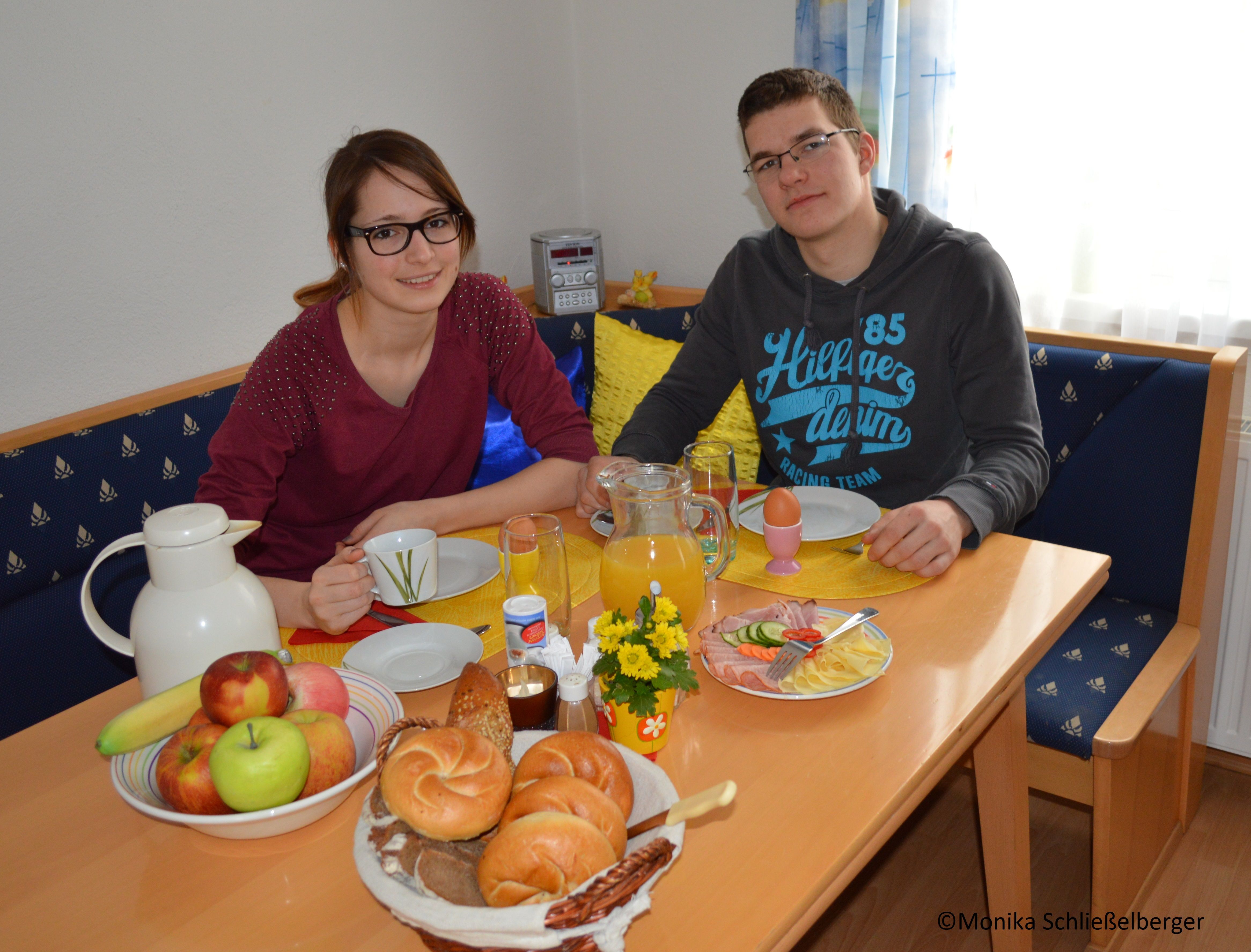 Zwei Personen sitzen an einem Tisch mit Frühstück, darunter Brötchen, Obst und Saft.