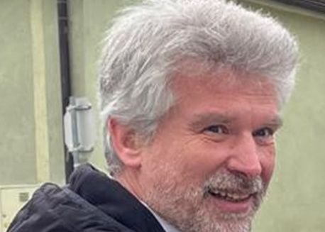 Ein lächelnder Mann mit grauem Haar und Bart vor einem grünen Gebäude.