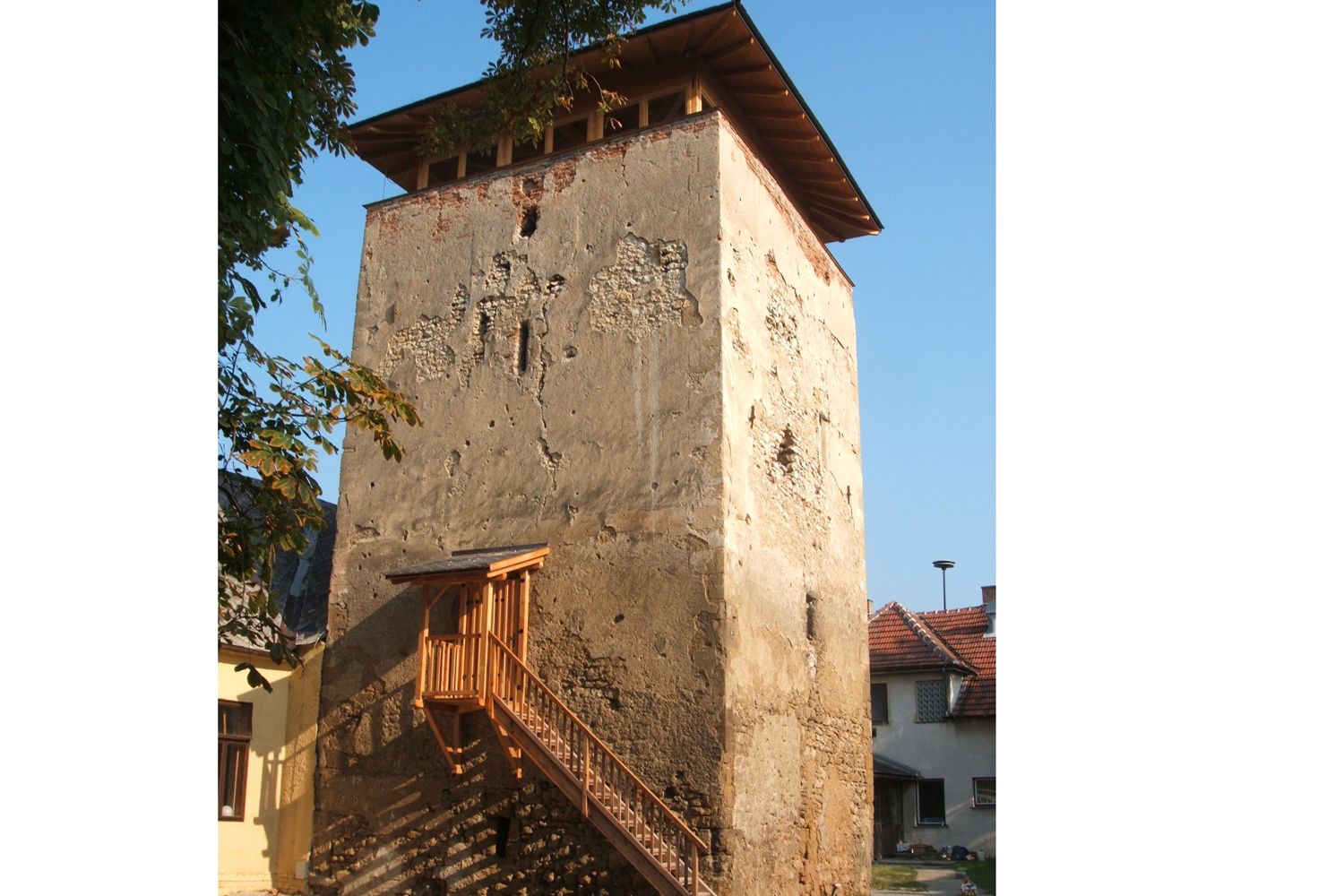Ein alter Wehrturm mit bröckelnder Fassade und einem hölzernen Treppenaufgang, umgeben von Häusern und Bäumen.
