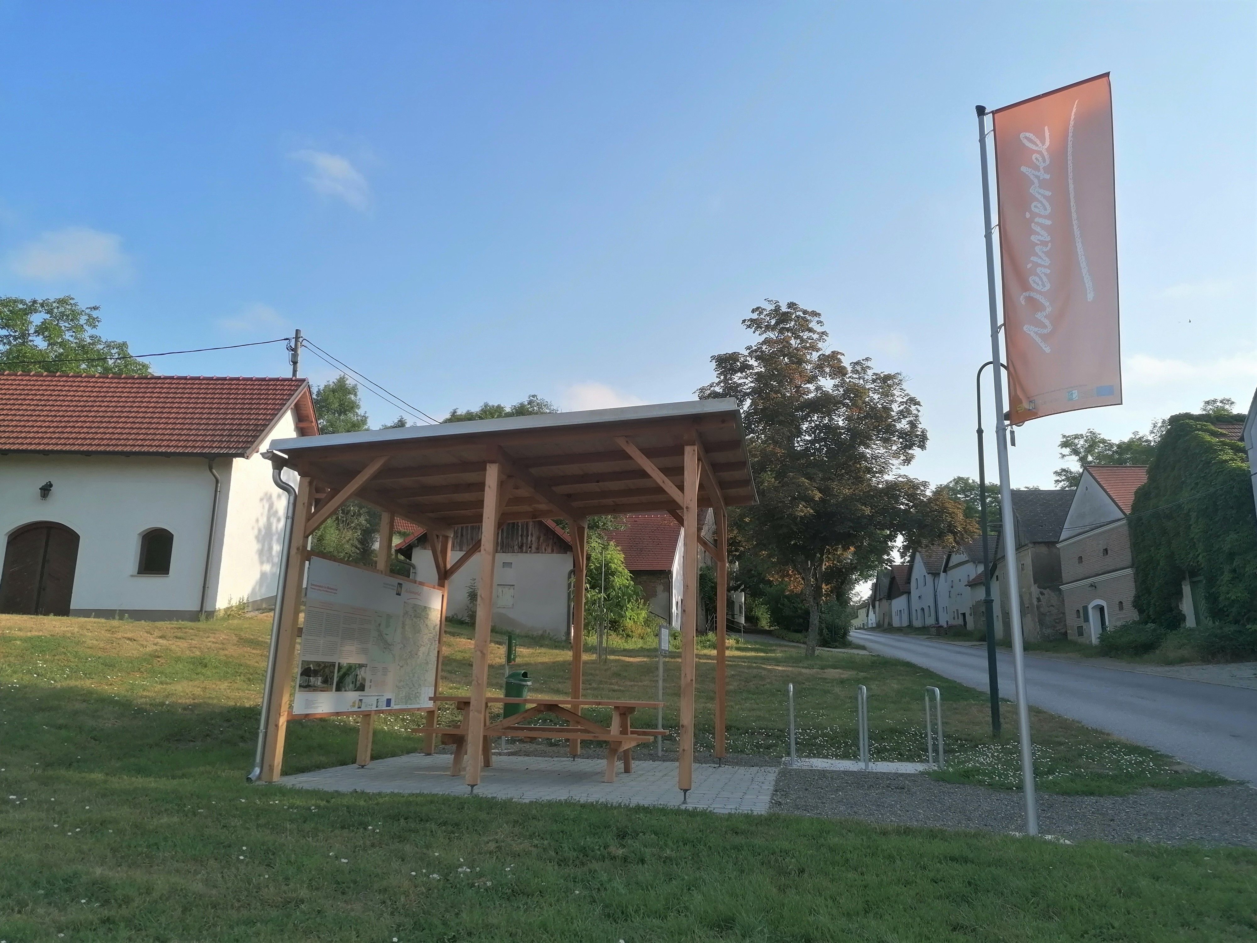 Ein Rastplatz mit Holzdach und Sitzbank neben einer Straße in Großkrut, umgeben von traditionellen Gebäuden und einer Fahne mit der Aufschrift 'Weinviertel'.
