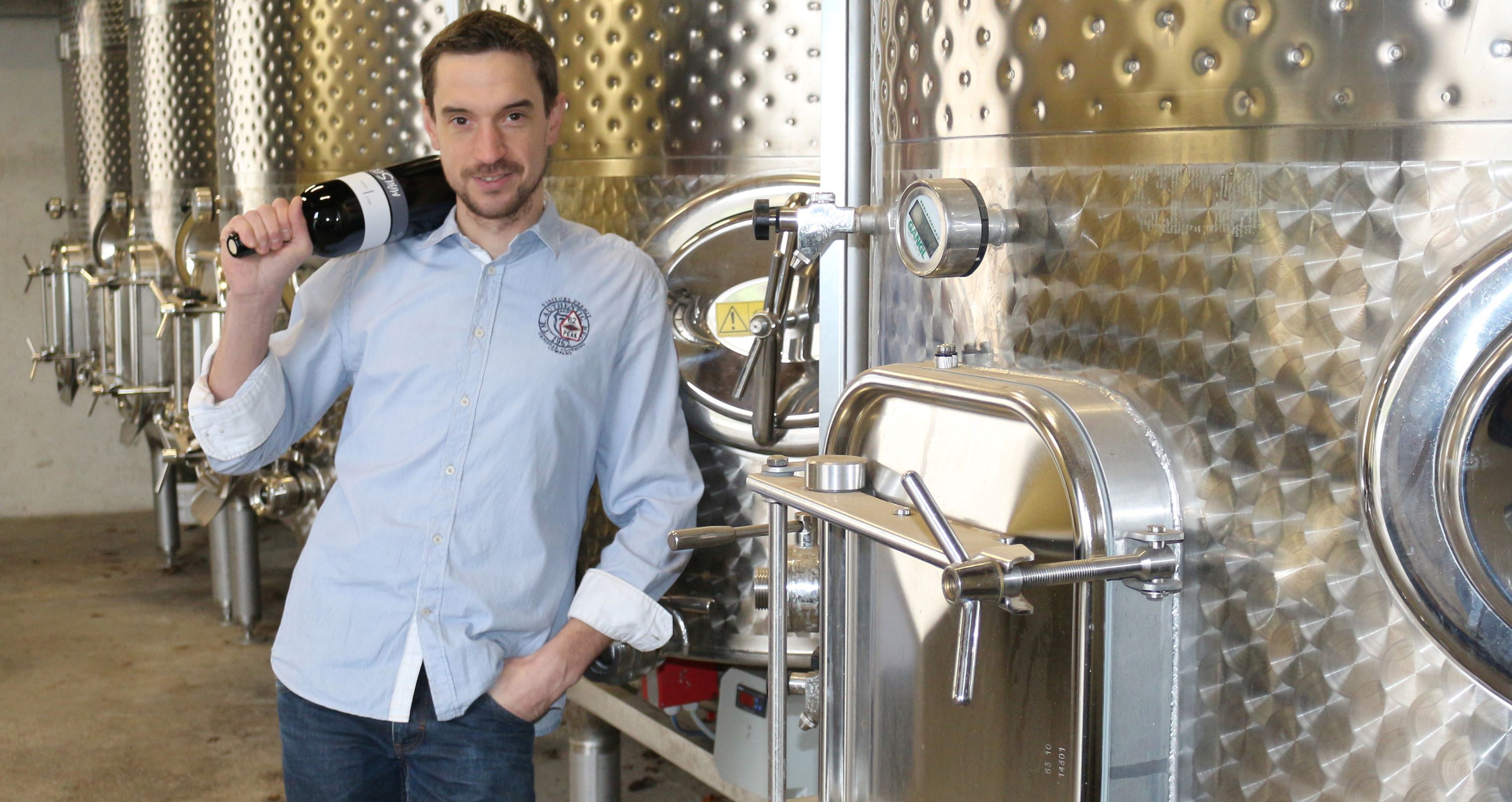 Ein Winzer steht vor Stahltanks in seinem Weinkeller, er hält lässig über der Schulter eine Flasche Wein in der Hand.