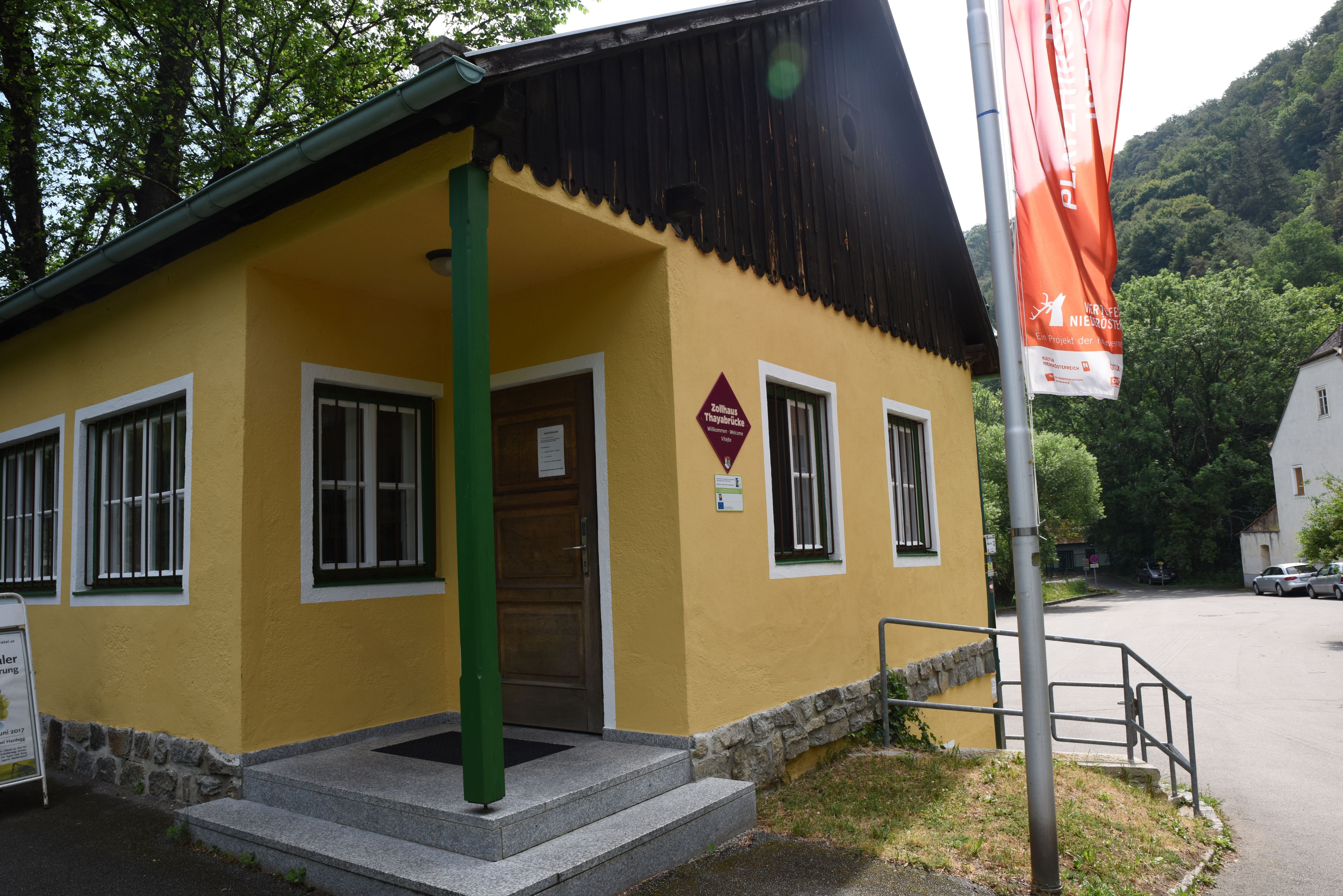 Das Zollhaus an der Thayabrücke in Hardegg mit gelber Fassade, grüner Stütze und roter Fahne vor bewaldetem Hang.