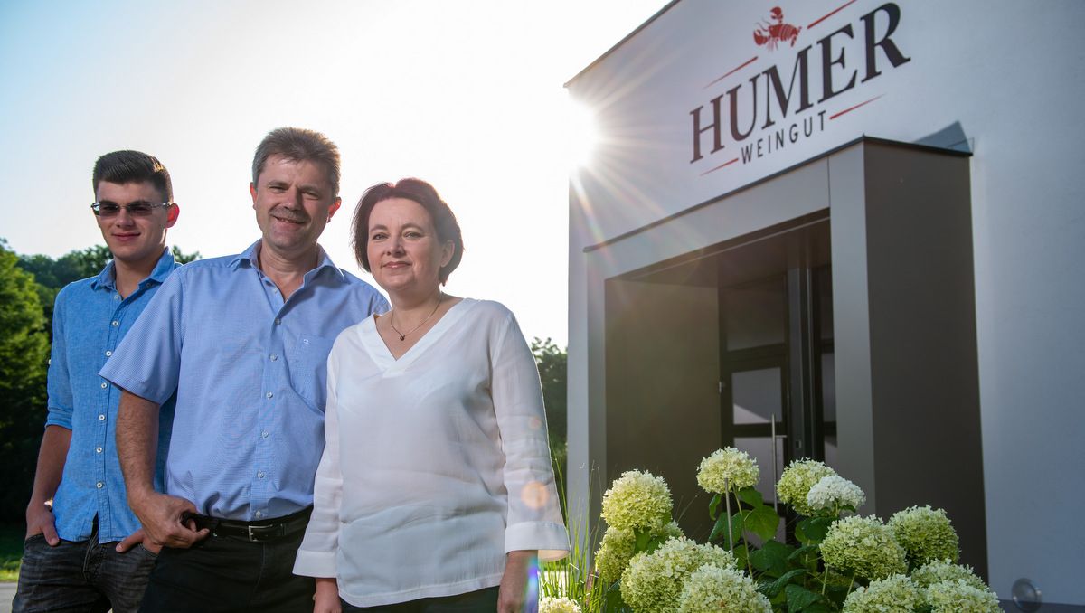 Drei Personen vor dem Eingang der Humer Weinkellerei, umgeben von Blumen.