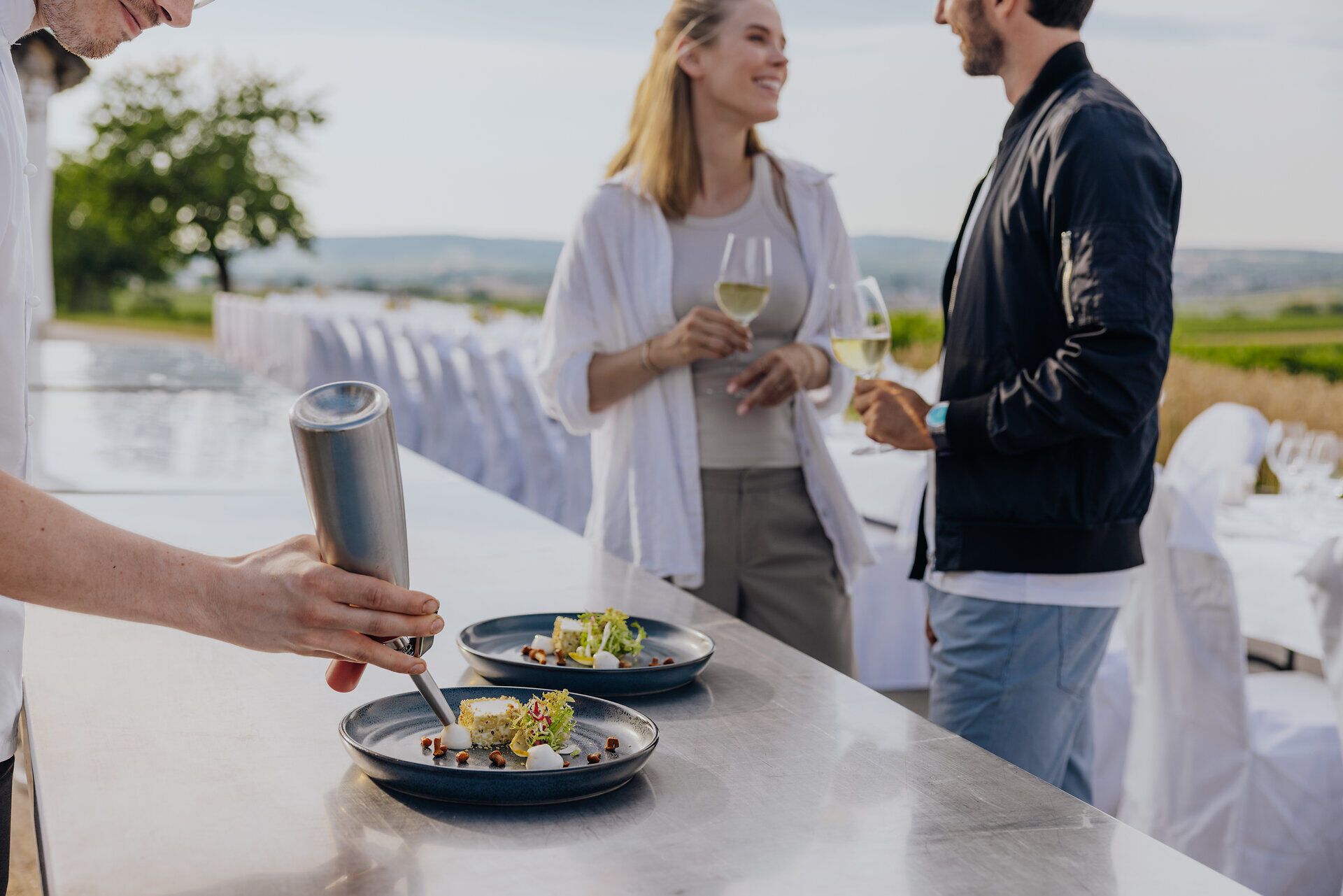 Inmitten der malerischen Weinlandschaft genießen Paare ein kulinarisches Erlebnis, während ein talentierter Koch kunstvoll ein Gericht anrichtet. Die sanften Hügel und die frische Luft schaffen eine einladende Atmosphäre, die zum Verweilen einlädt.