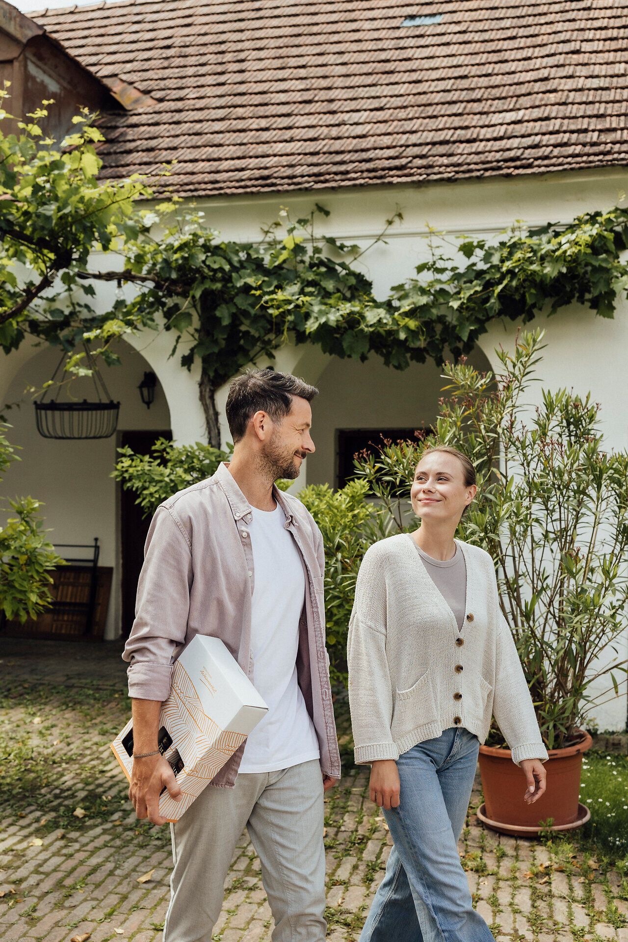 Ein Paar schlendert entspannt durch die malerische Umgebung eines Winzerhofs, während sie einen Weinkarton in der Hand halten. Die sanften Hügel und üppigen Weinreben schaffen eine einladende Atmosphäre, die zum Genießen und Verweilen einlädt.