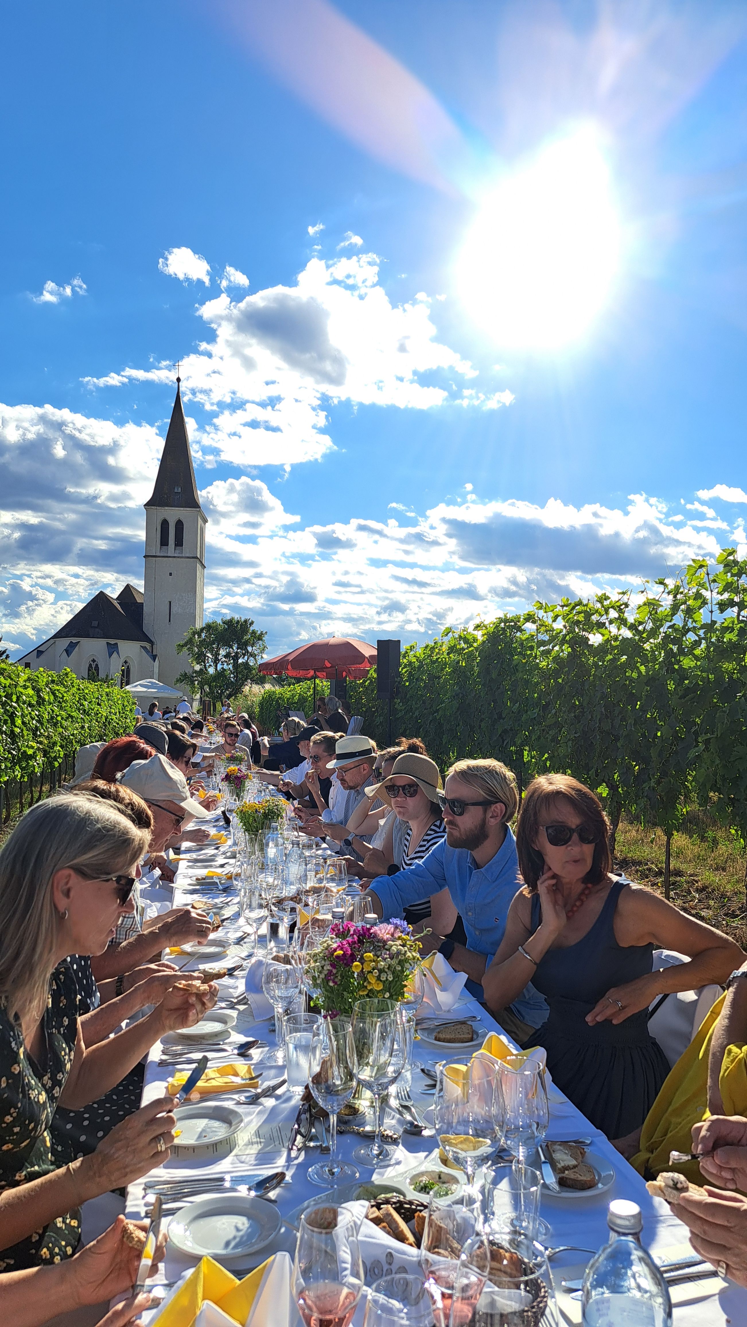 Menschen sitzen an einer langen Tafel im Freien neben einem Weinberg, mit einer Kirche im Hintergrund und strahlendem Sonnenschein.