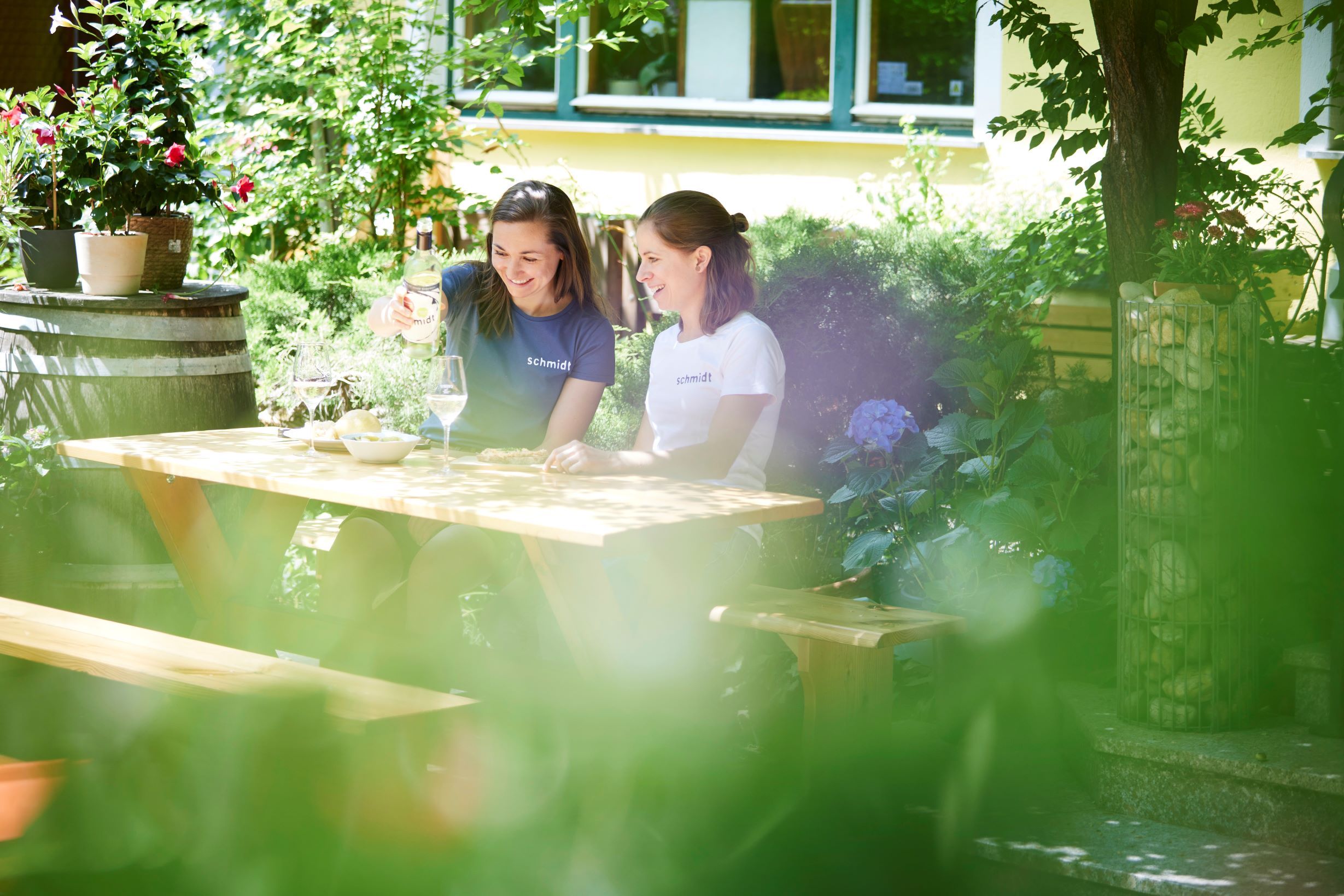 Zwei Frauen sitzen an einem Holztisch im Garten und genießen Wein.