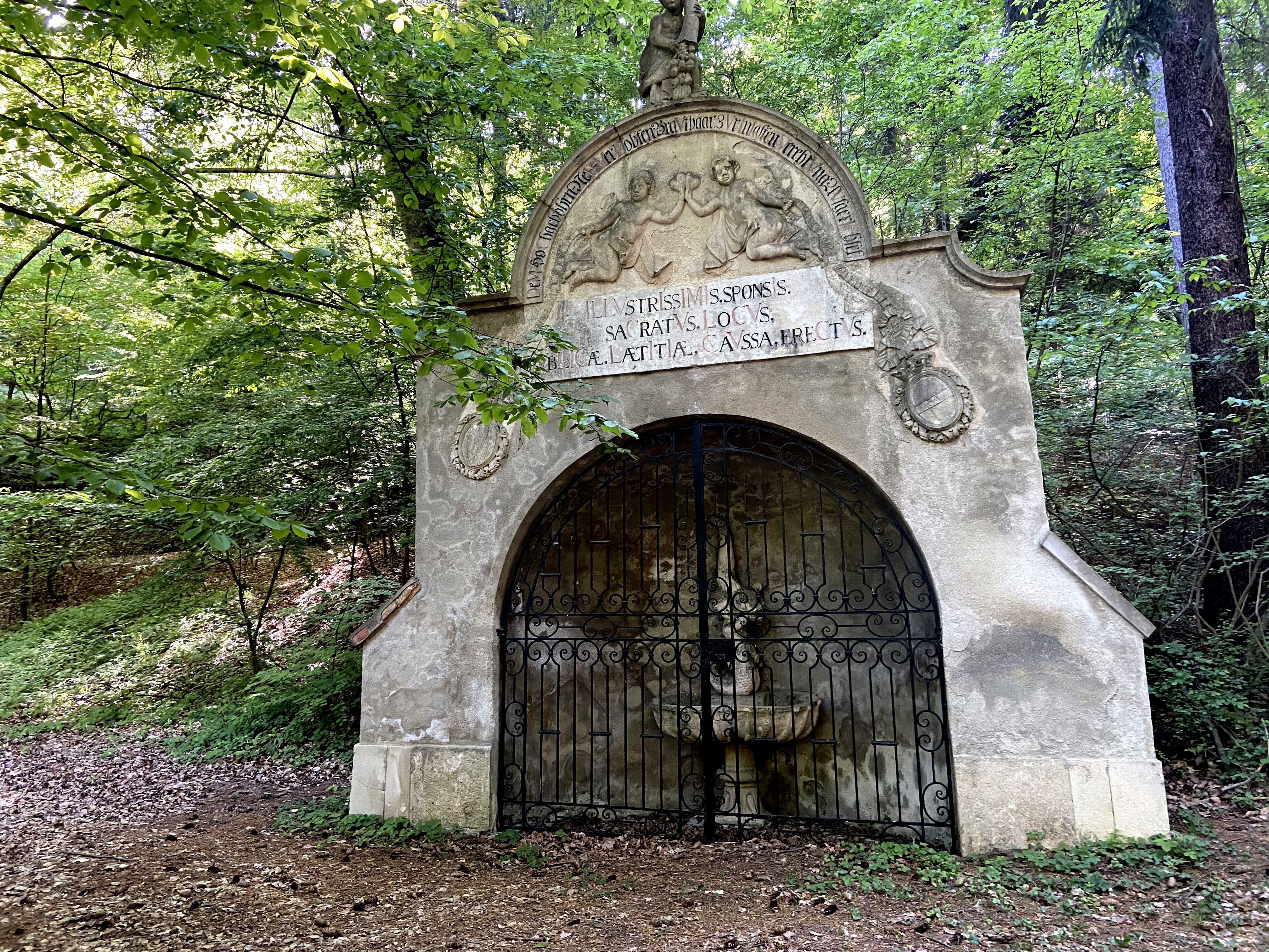 Historische Brunnstube im Wald mit verziertem Tor und lateinischer Inschrift.