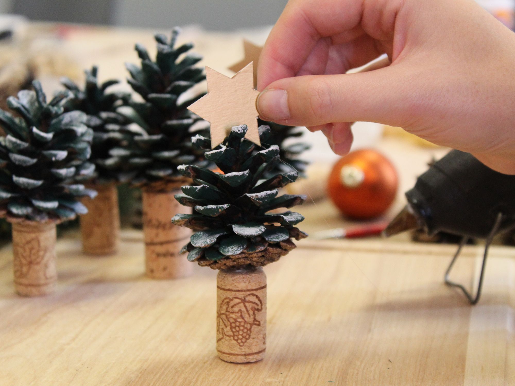 Ein Tannenzapfen steht auf einem Korken und ein goldener Stern wird als Spitze darauf geklebt. Im Hintergrund stehen noch mehr Tannenbäumchen.