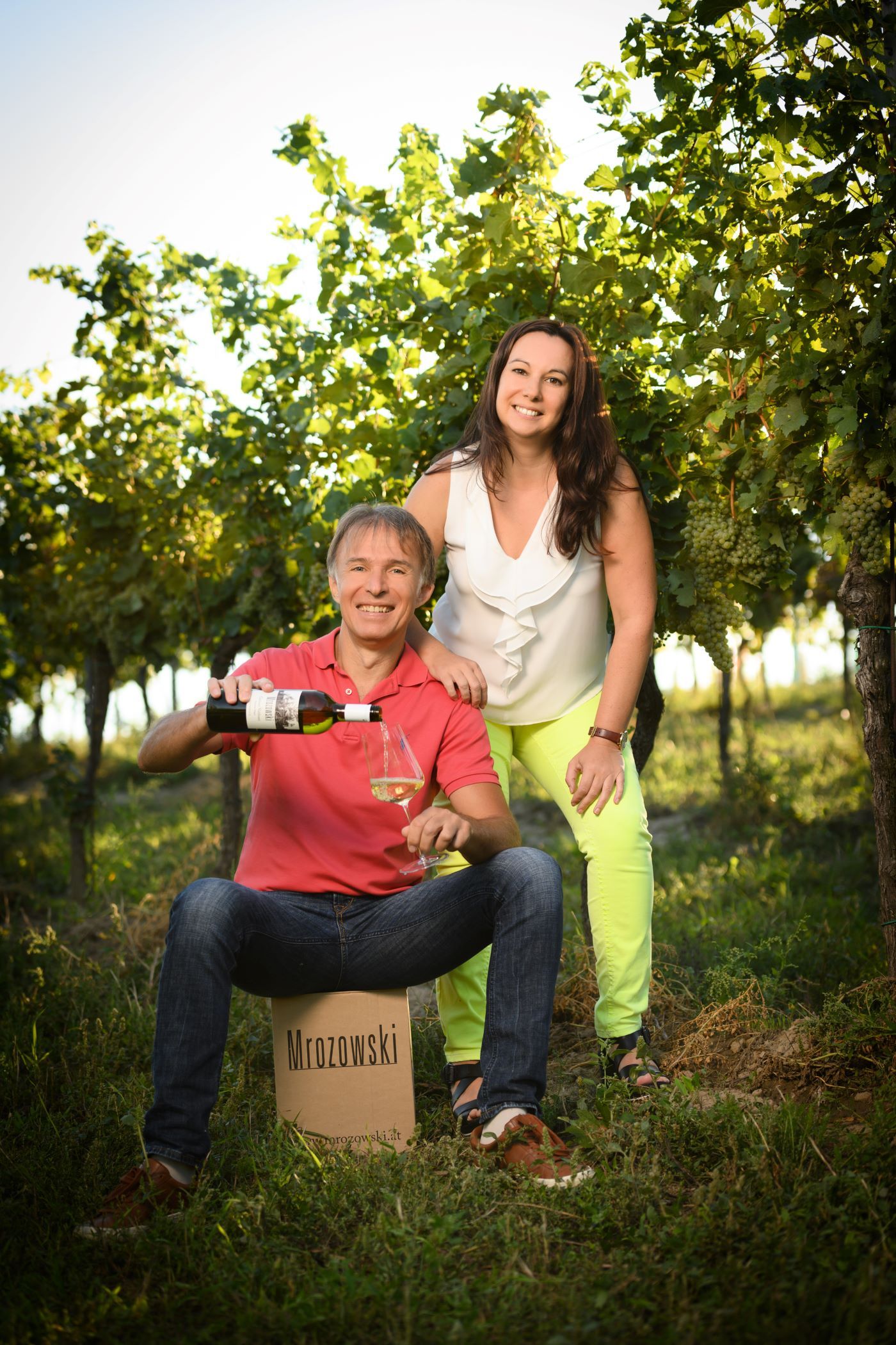 Two people in a vineyard, one sits on a crate and pours wine, the other stands next to it and smiles.
