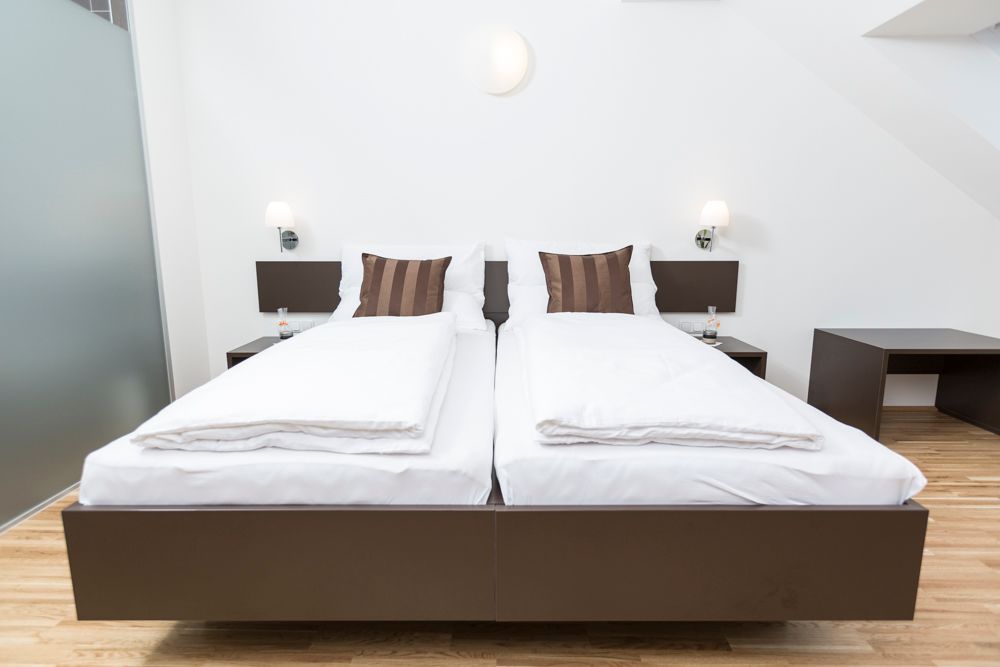 Modernes Gästezimmer mit zwei Einzelbetten, weißen Bettdecken und braunen Kissen, Holzboden und Nachttischen mit Lampen.