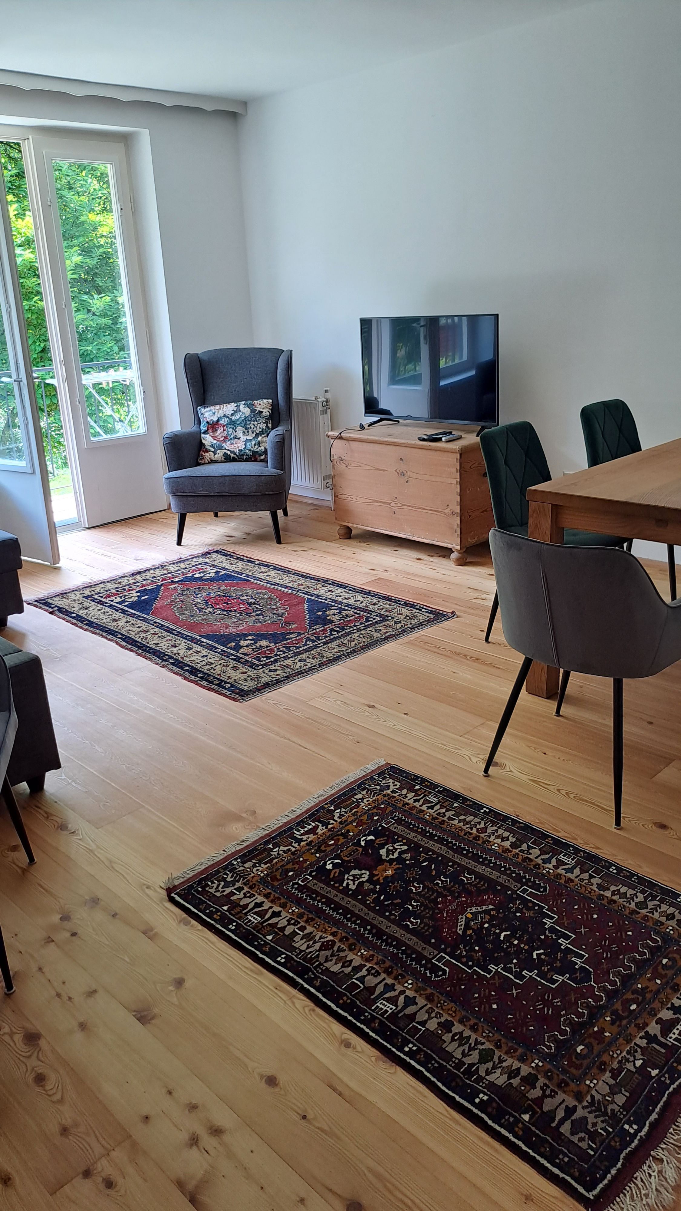 Wohnzimmer mit Holzboden, Teppichen, einem Sessel, einem Fernseher und einem Esstisch mit Stühlen.