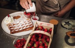 Ein Korb voller Erdbeeren und eine frische Waffel auf einem Teller, eine Hand gießt Erdbeersauce in ein kleines Gefäß.