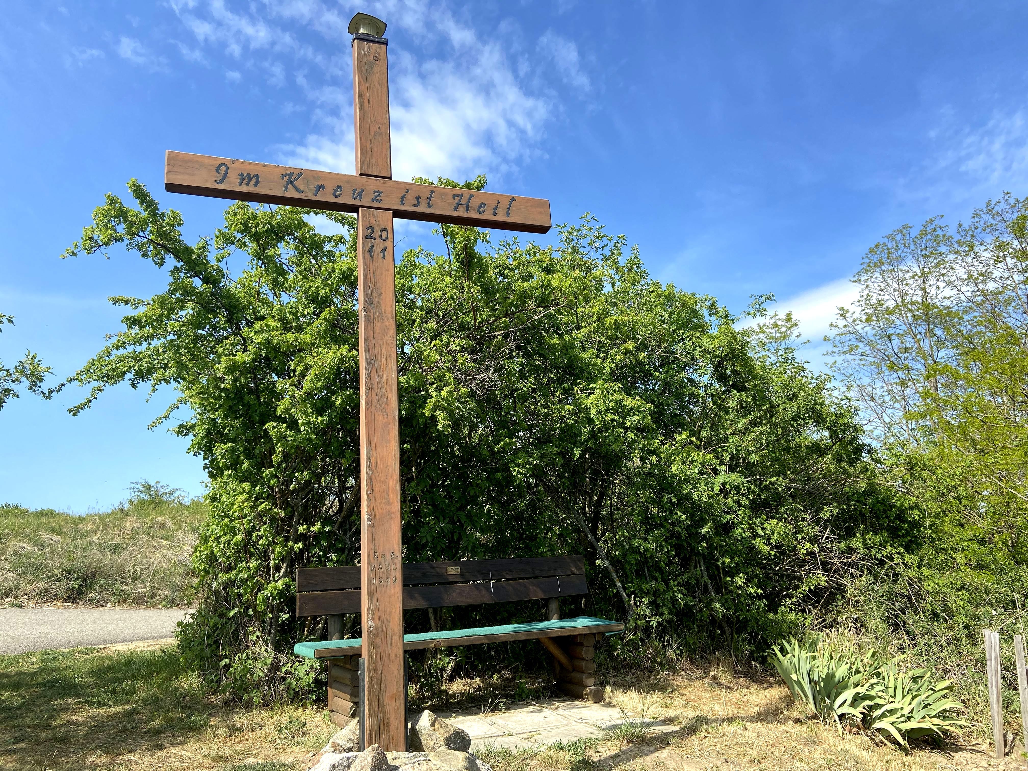 Holzkreuz mit Inschrift 'Im Kreuz ist Heil' und Sitzbank, umgeben von Büschen und blauem Himmel.