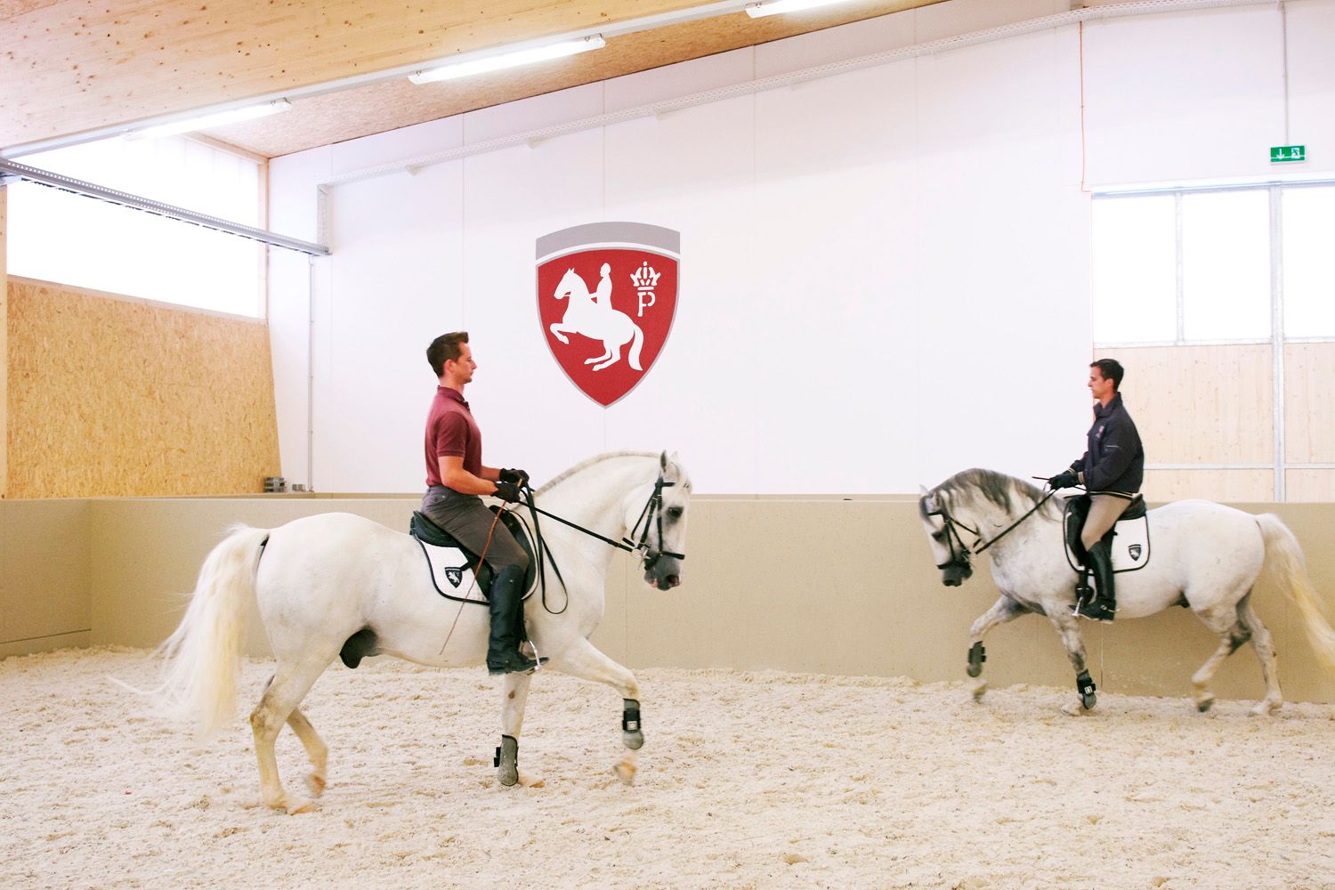 Zwei Reiter auf Lipizzanern in einer Reithalle mit einem Wappen an der Wand.