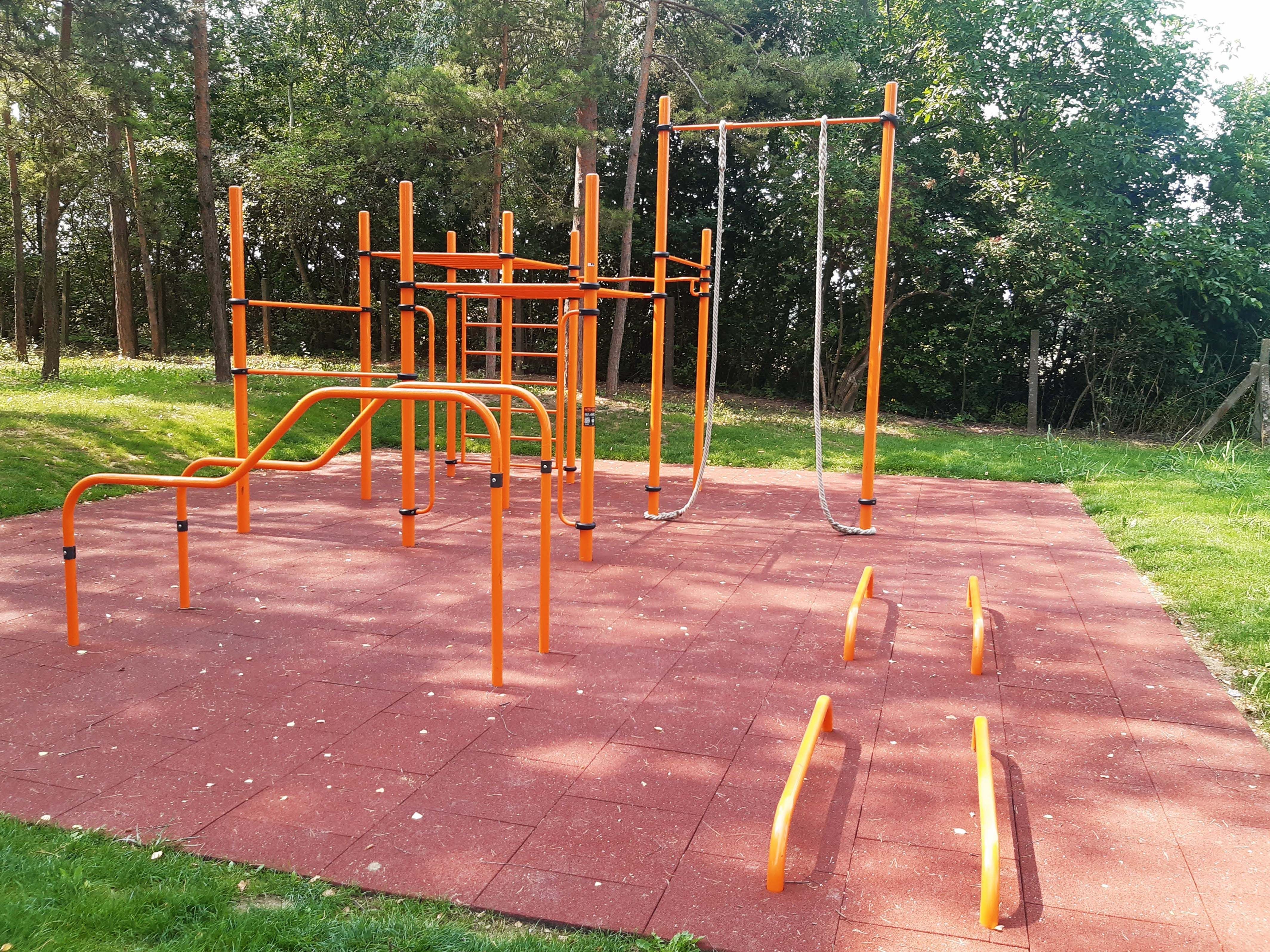 An outdoor fitness park with orange climbing frames and poles on a red rubber floor, surrounded by trees and grass.