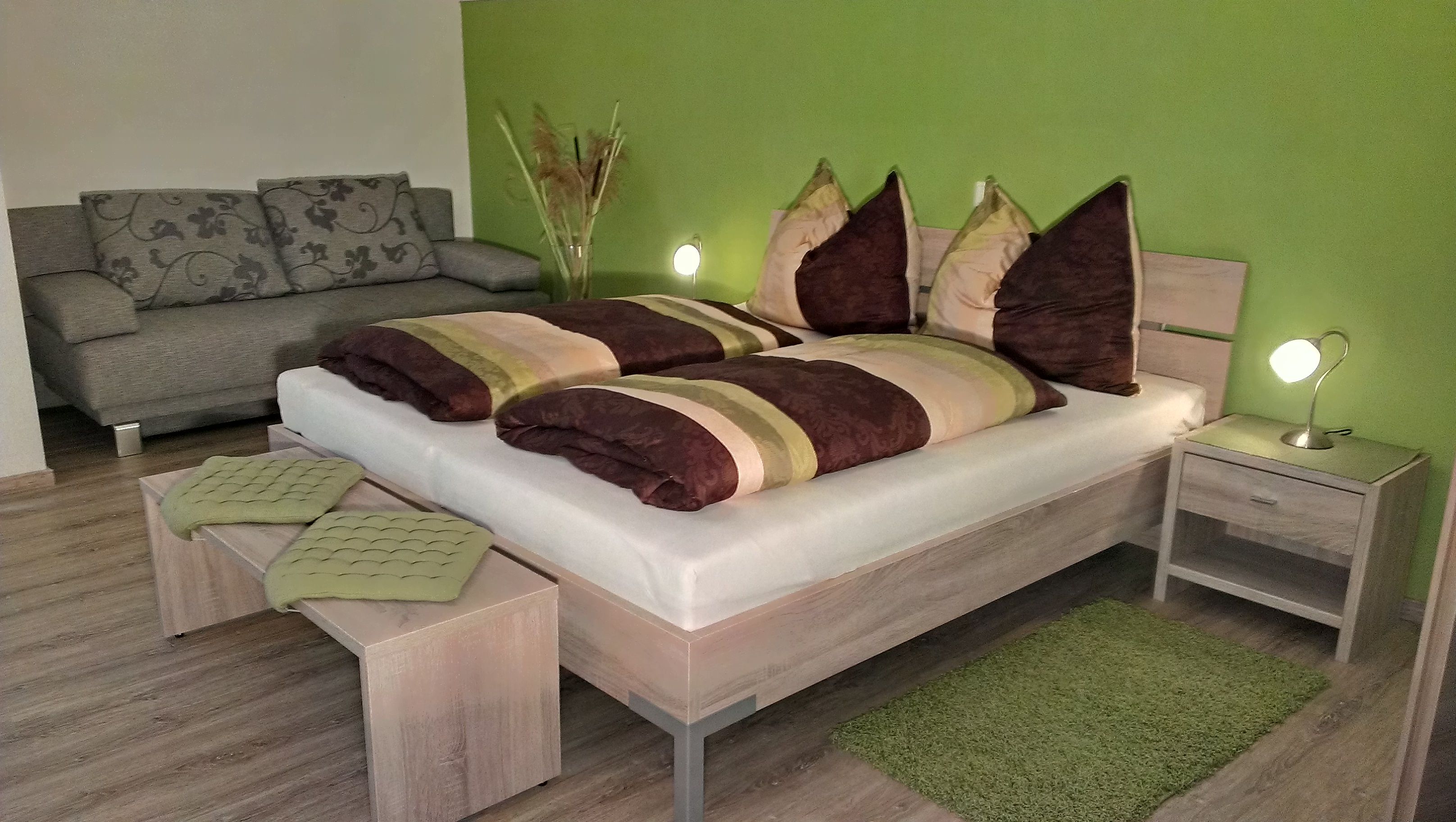 Gemütliches Schlafzimmer mit grün gestrichener Wand, Doppelbett, Nachttischlampen und grauem Sofa.