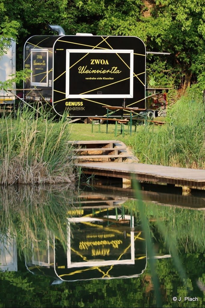 Ein schwarzer Foodtruck mit der Aufschrift 'ZWOA Weinviertla' steht in einer grünen Umgebung, reflektiert im Wasser.
