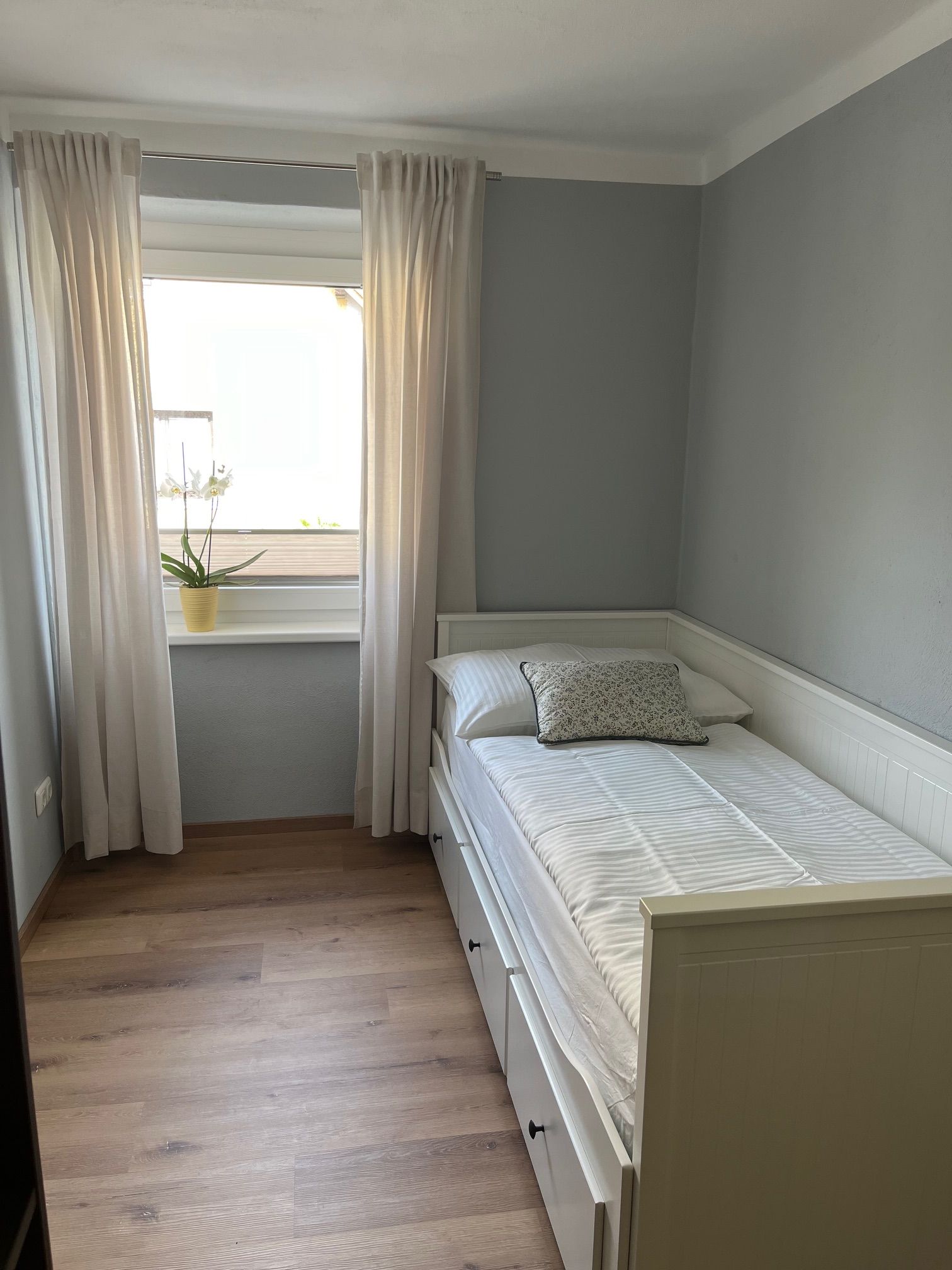 Kleines Schlafzimmer mit Einzelbett, weißen Vorhängen und einer Pflanze auf der Fensterbank.