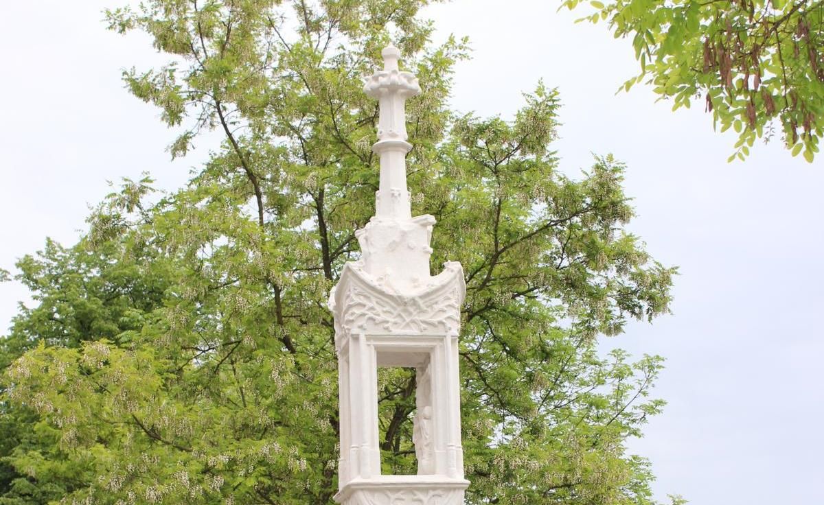 Weiße Steinsäule mit Kreuz vor grünem Baum.