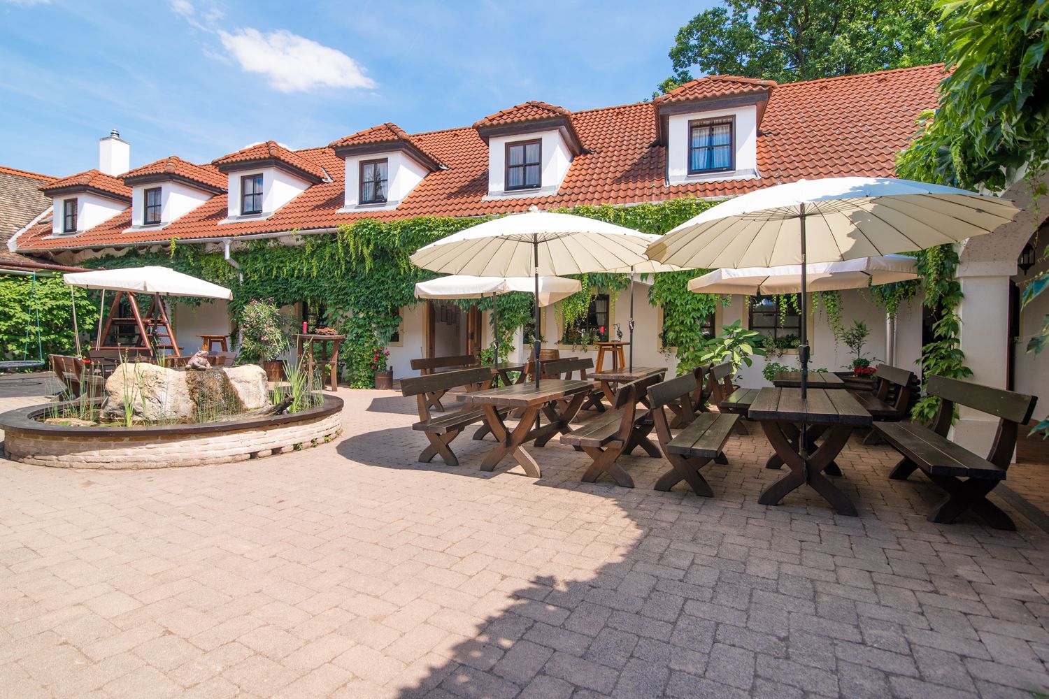 Ein gemütlicher Innenhof eines Weinguts mit Holztischen, Bänken und Sonnenschirmen, umgeben von einem Gebäude mit roten Ziegeldächern und grüner Bewachsung.