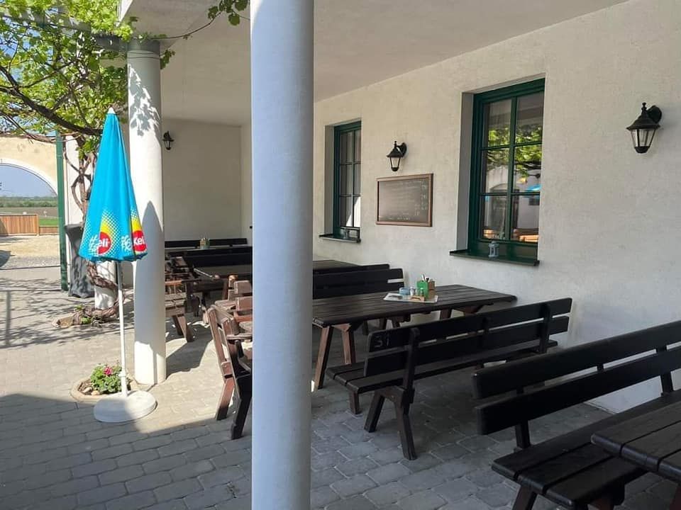 Ein leerer Gastgarten mit Holzbänken und Tischen, einem Sonnenschirm und einer weißen Wand mit Fenstern.