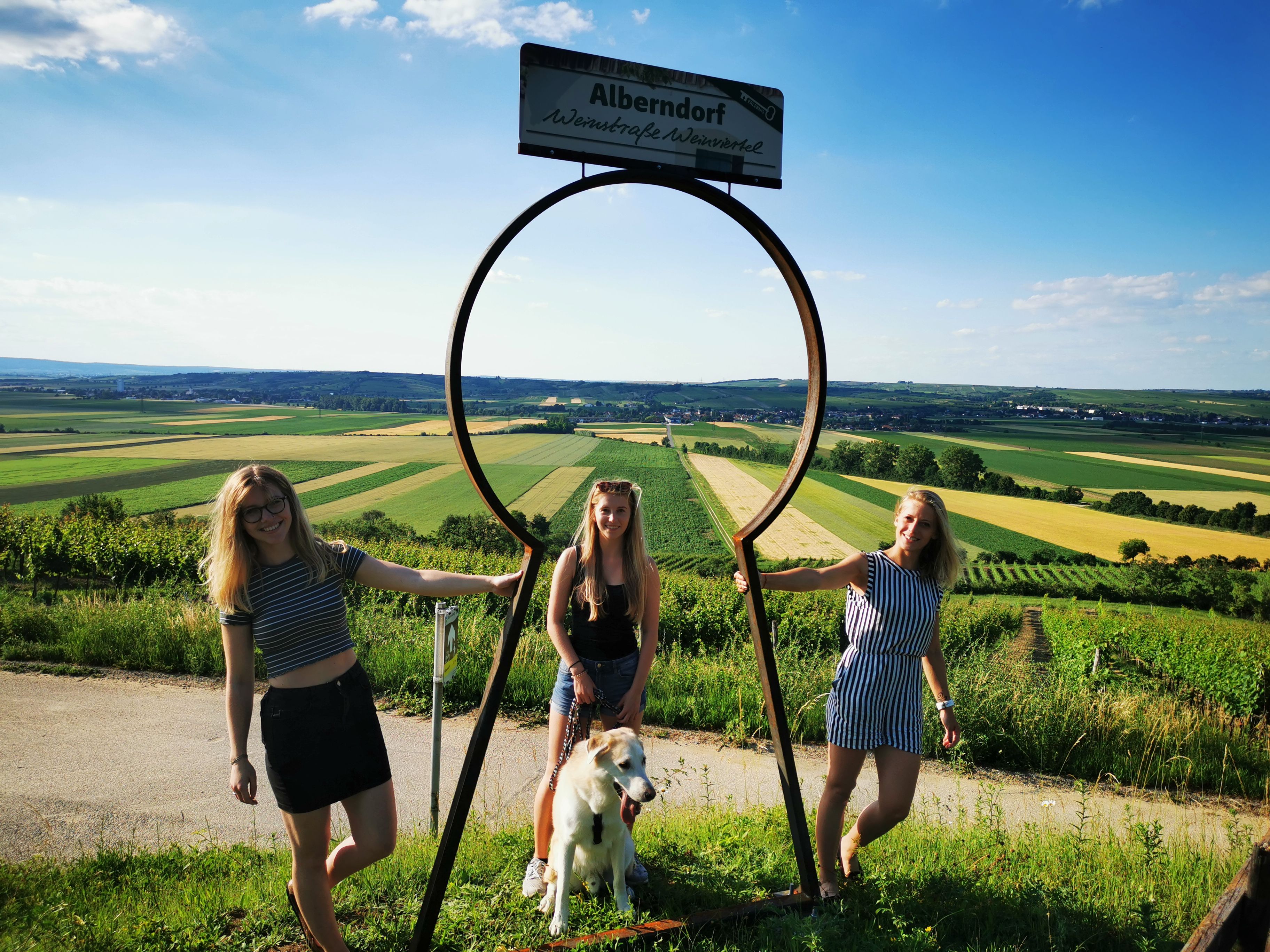 Drei Frauen und ein Hund posieren vor einem großen Schlüssellochrahmen mit Blick auf eine ländliche Landschaft.