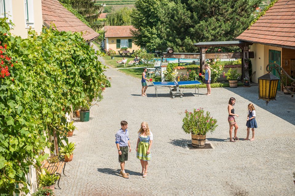 Ein sonniger Hof mit Menschen, die spazieren, Tischtennis spielen und sich unterhalten. Umgeben von Pflanzen und Gebäuden mit roten Dächern.