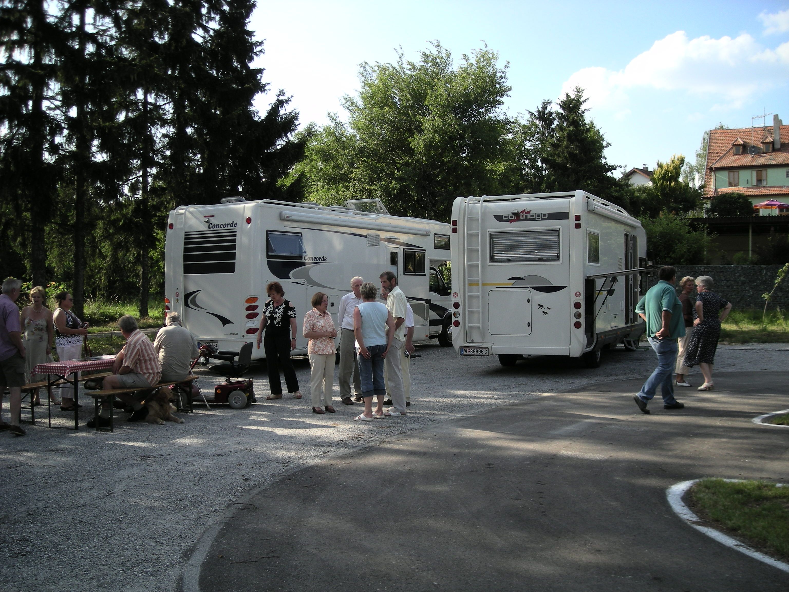 Menschen stehen und sitzen neben zwei Wohnmobilen auf einem Stellplatz im Freien.