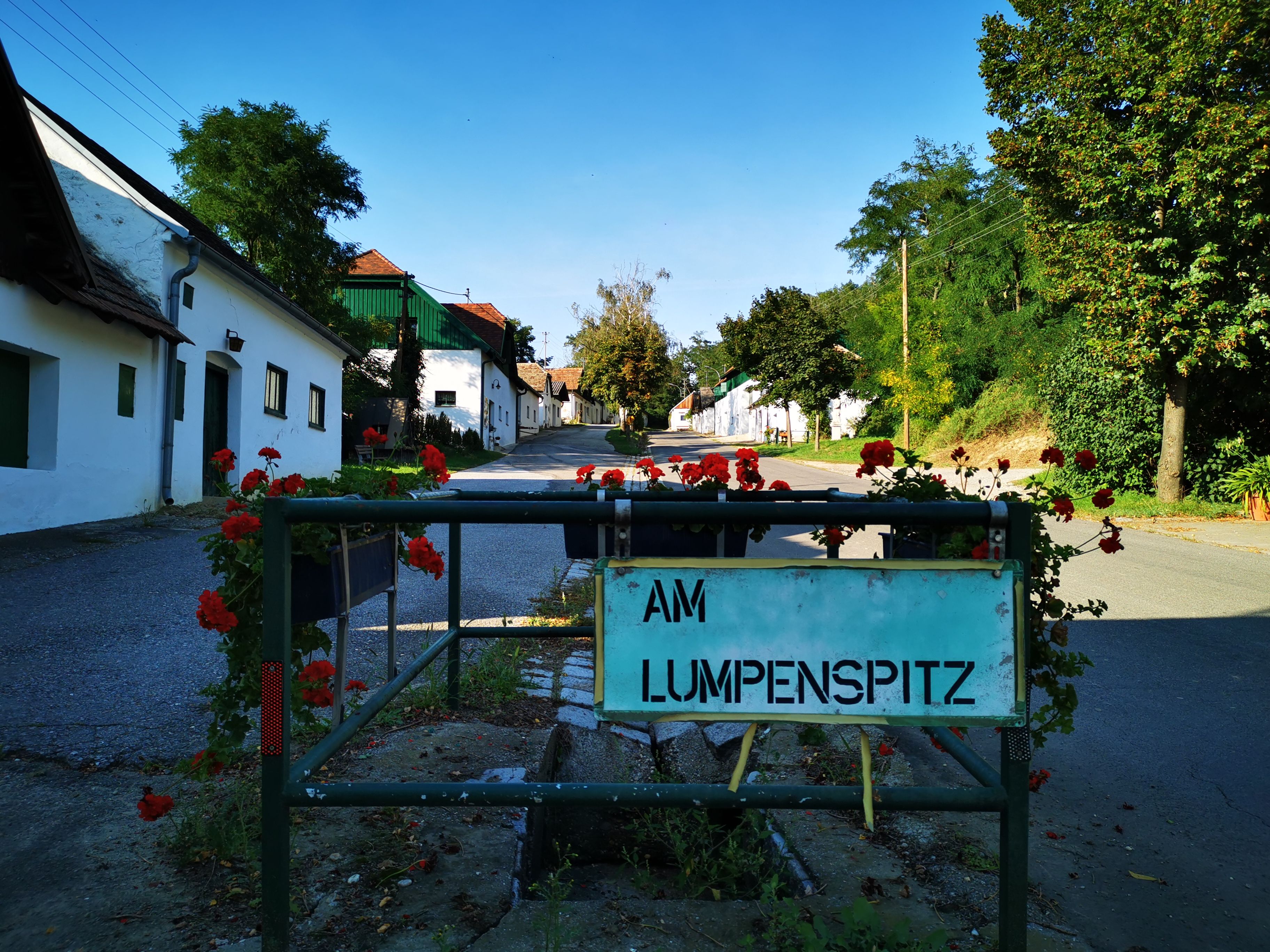 Straßenschild 'Am Lumpenspitz' in einer malerischen Kellergasse mit blühenden Geranien und traditionellen Gebäuden.