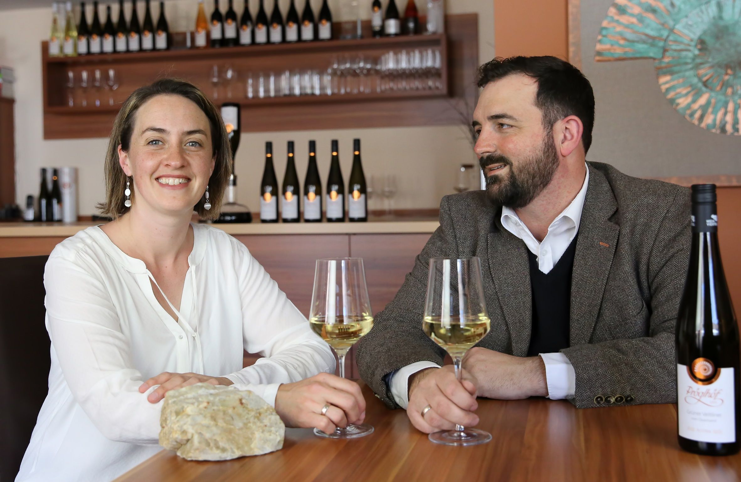 Zwei Personen sitzen an einem Tisch mit Weingläsern und Weinflaschen im Hintergrund.