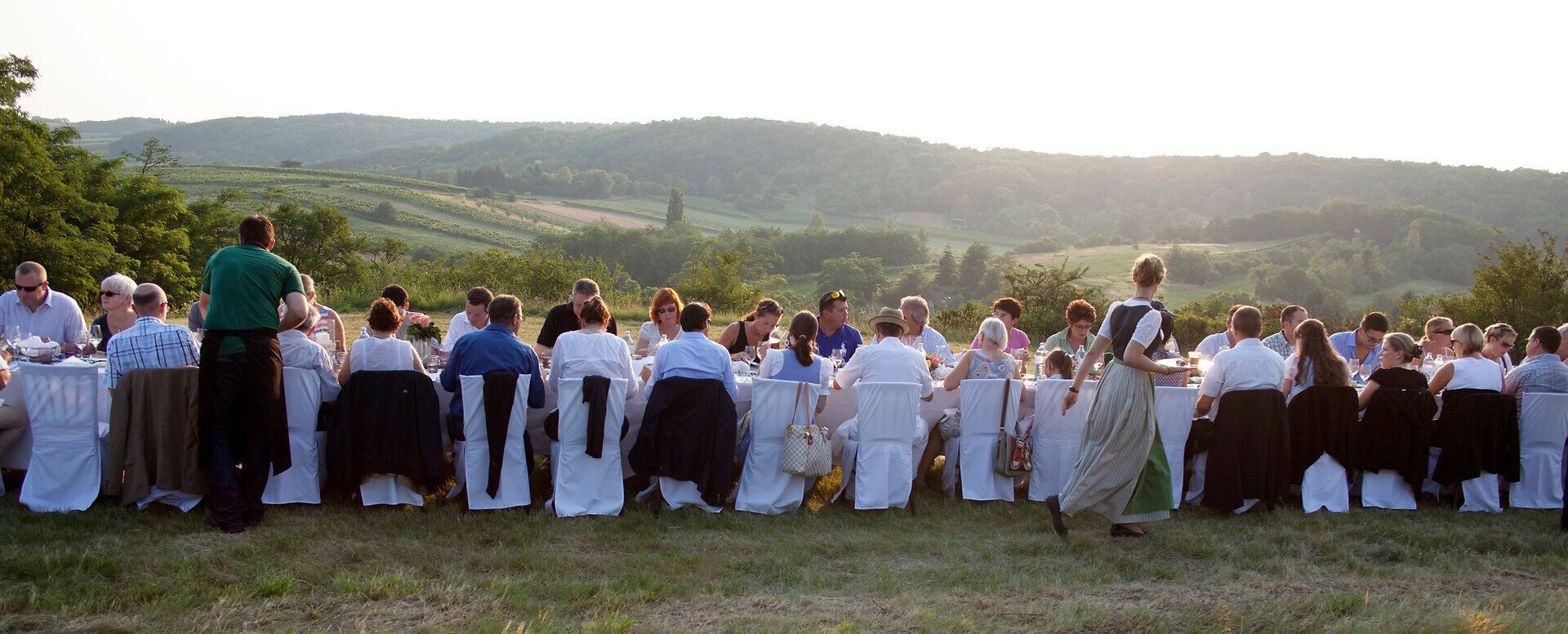 Eine lange Tafel, festlich gedeckt, zieht sich durch die malerische Landschaft, während die Sonne sanft hinter den Hügeln untergeht. Die Gäste genießen die köstlichen Speisen und die gesellige Atmosphäre, umgeben von der Schönheit der Natur. Ein unvergesslicher Abend, der die Sinne verzaubert.