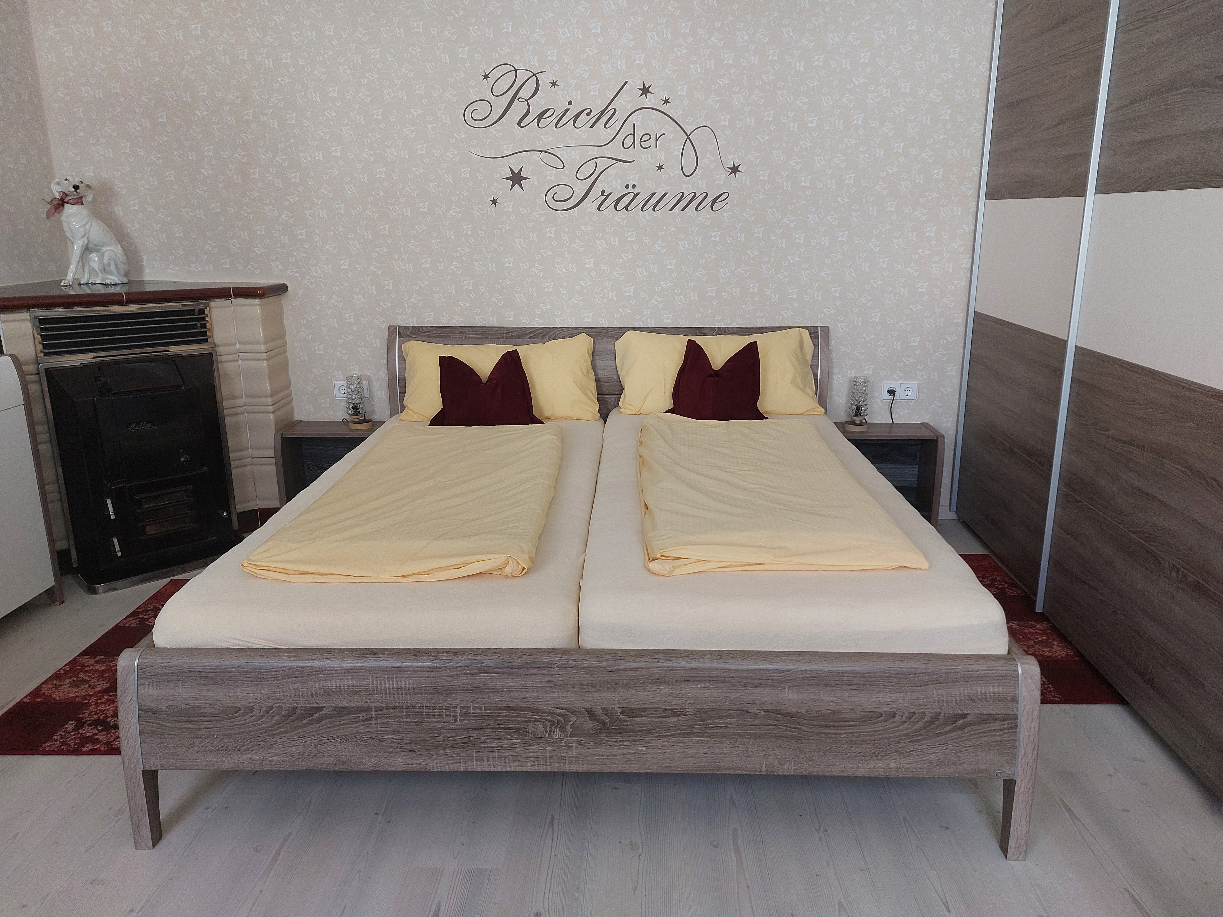 Ein Schlafzimmer mit einem Doppelbett, gelber Bettwäsche und einem Schriftzug an der Wand. Ein Ofen und eine Hundeskulptur sind ebenfalls zu sehen.