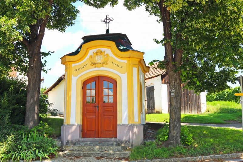 Kleine gelbe Kapelle mit Holztür, umgeben von Bäumen und Grünflächen.
