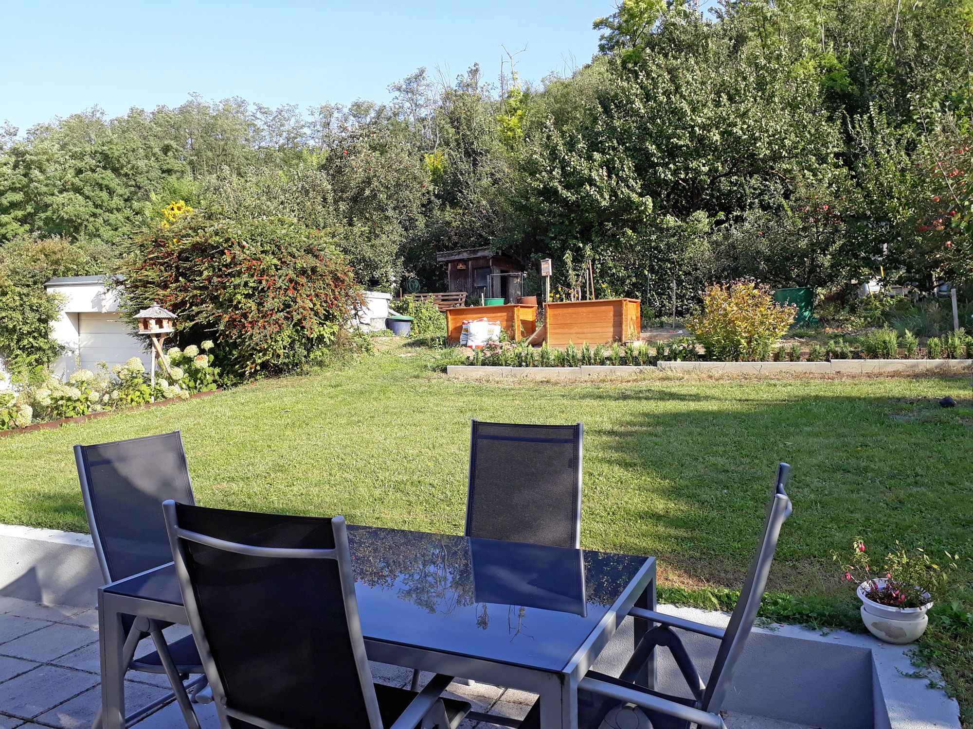 Terrasse mit Tisch und Stühlen, Blick auf einen Garten mit Rasen, Bäumen und Hochbeeten.
