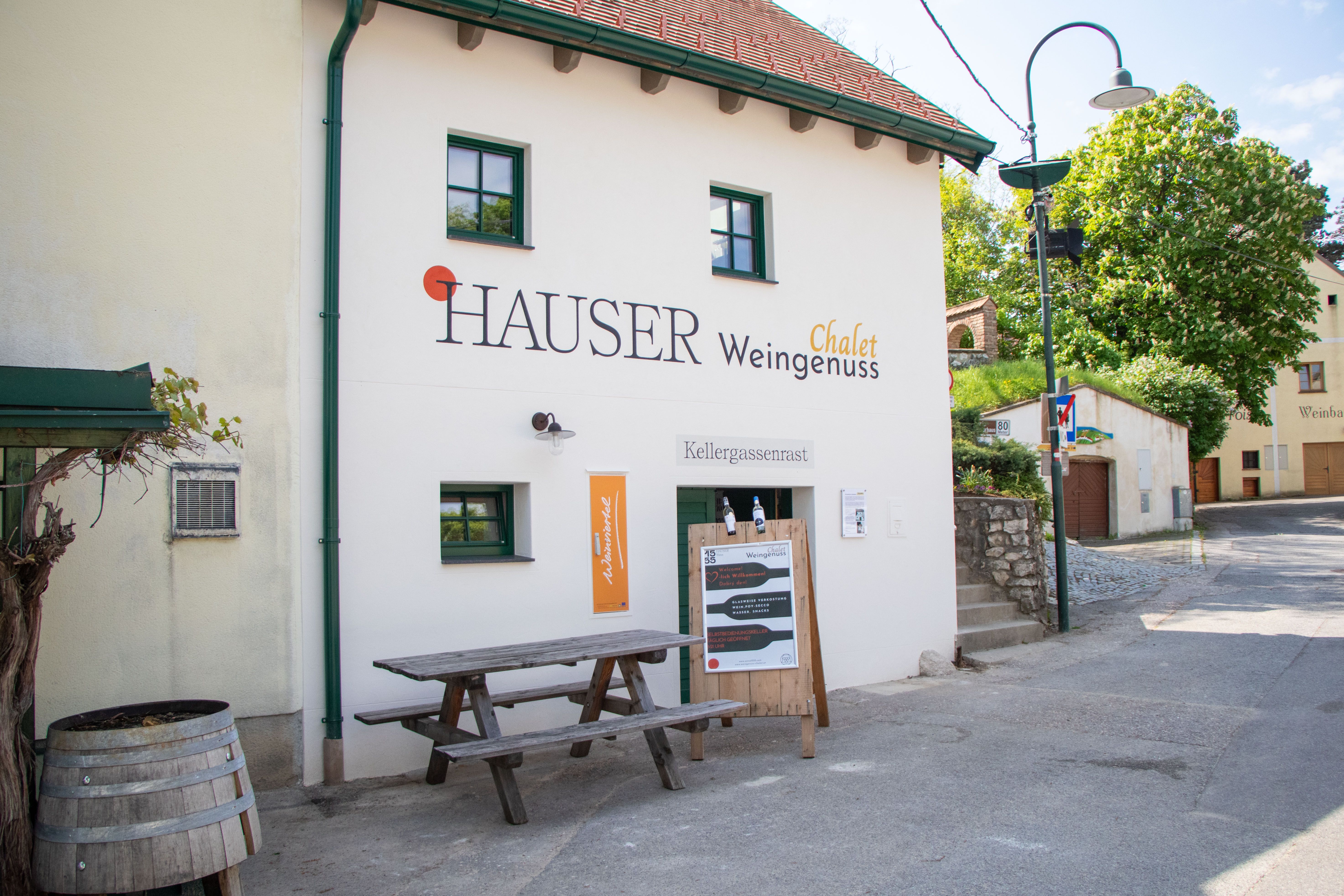 Exterior view of a chalet with the inscription 'Hauser Weingenuss'. In front of the building is a wooden table and a board with information.