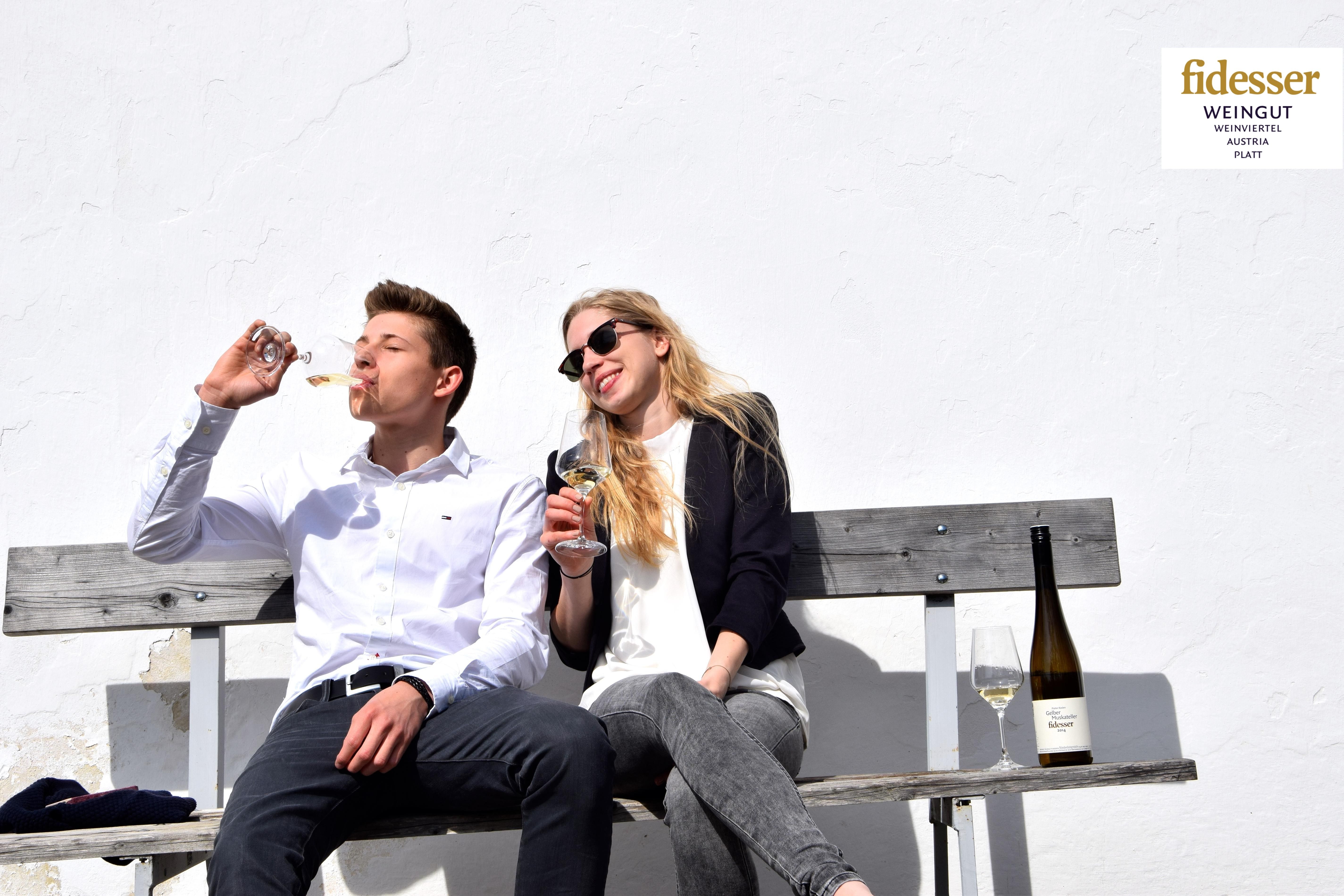 Zwei Personen sitzen auf einer Bank und trinken Wein vor einer weißen Wand.