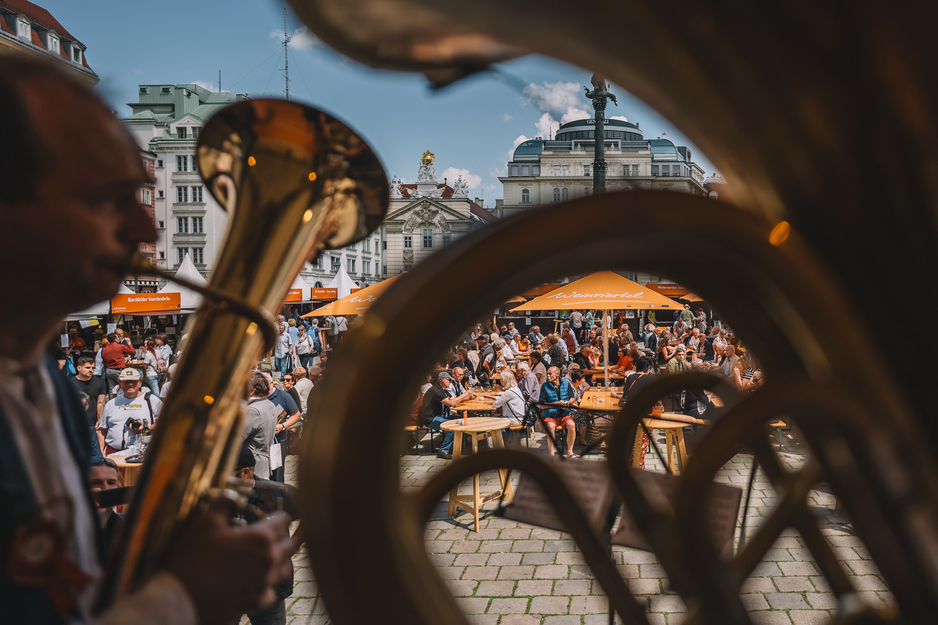 Durch zwei Musikanten, welche auf der Bühne sitzen und musizieren, sieht man Personen, welche Weinviertler Getränke und Kulinarik in einer Innenstadt bei Sonnenschein genießen.