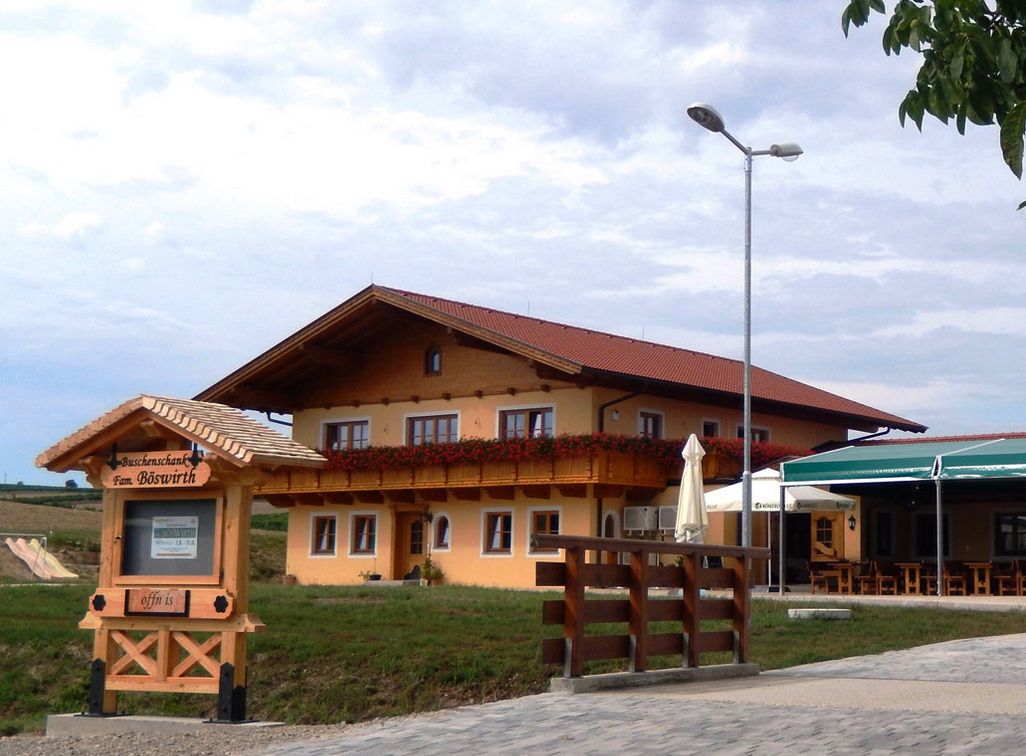 Ein traditionelles Gasthaus mit Holzbalkon und Blumendekoration, daneben ein Schild mit der Aufschrift 'Buschenschank Fam. Böswirth'.