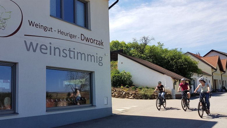 Drei Personen fahren mit Fahrrädern an einem Gebäude mit der Aufschrift 'Weingut - Heuriger - Dworzak weinstimmig' vorbei.