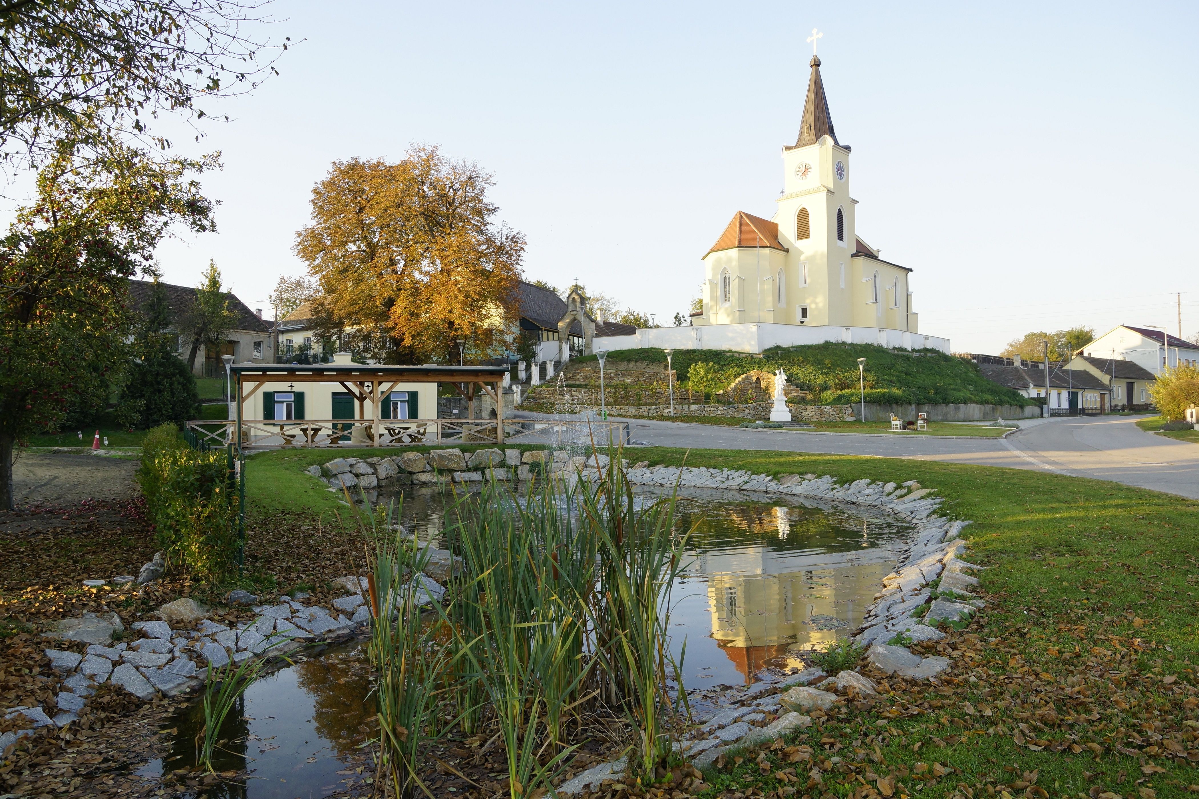 Dorfplatz in Glaubendorf mit Kirche, Teich und Herbstbäumen.