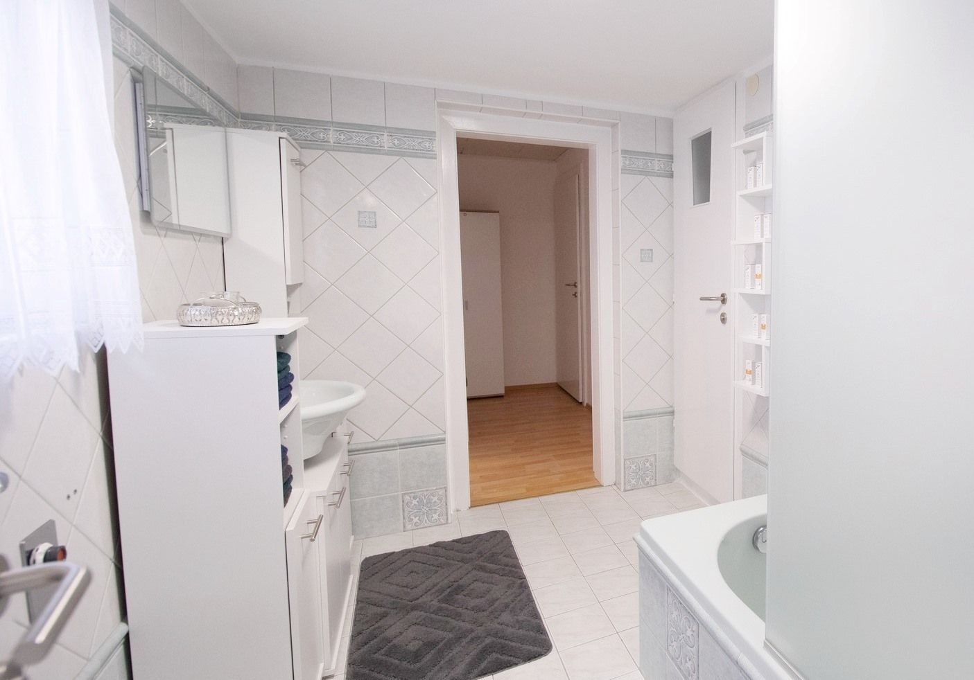 Modernes Badezimmer mit weißen Fliesen, Badewanne, Waschbecken und Schrank. Tür führt zu einem Flur mit Holzboden.