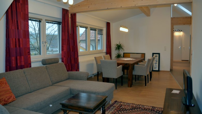 Wohnzimmer mit grauem Sofa, Holztisch und Stühlen, roten Vorhängen und Deckenleuchten.