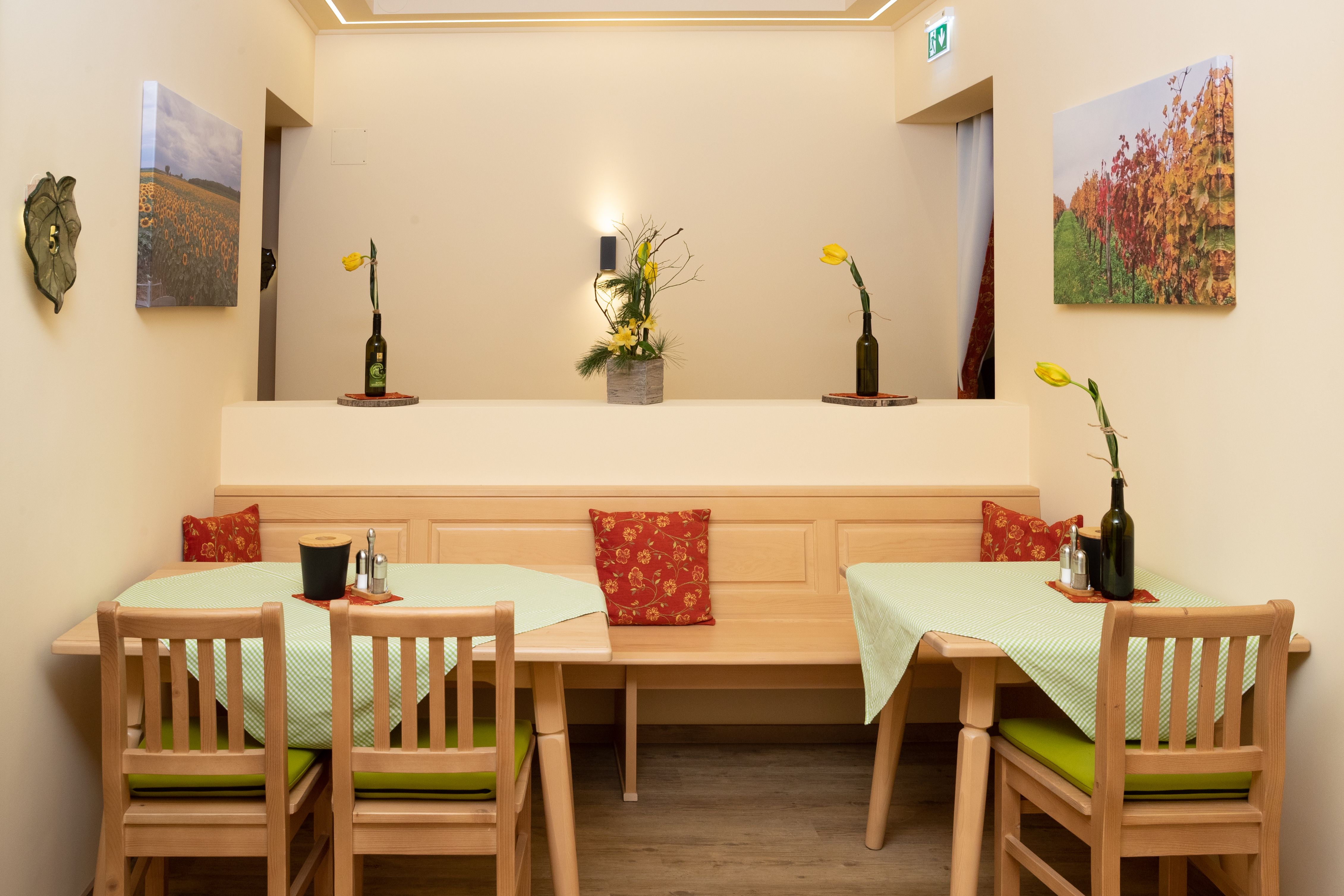 Ein gemütlicher Frühstücksraum mit Holzmöbeln, grünen Tischdecken und gelben Blumen in Flaschenvasen. An den Wänden hängen Landschaftsbilder.