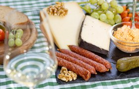 Ein gedeckter Tisch mit Käse, Wurst, Brot, Weintrauben und einem Glas Weißwein auf einer grün-weiß karierten Tischdecke.