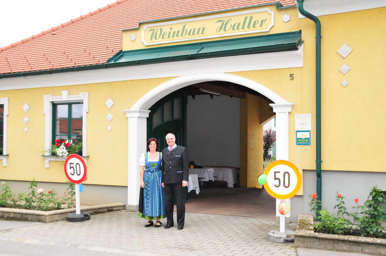 A couple is standing in front of a yellow building with the sign 'Weinbau Haller'.