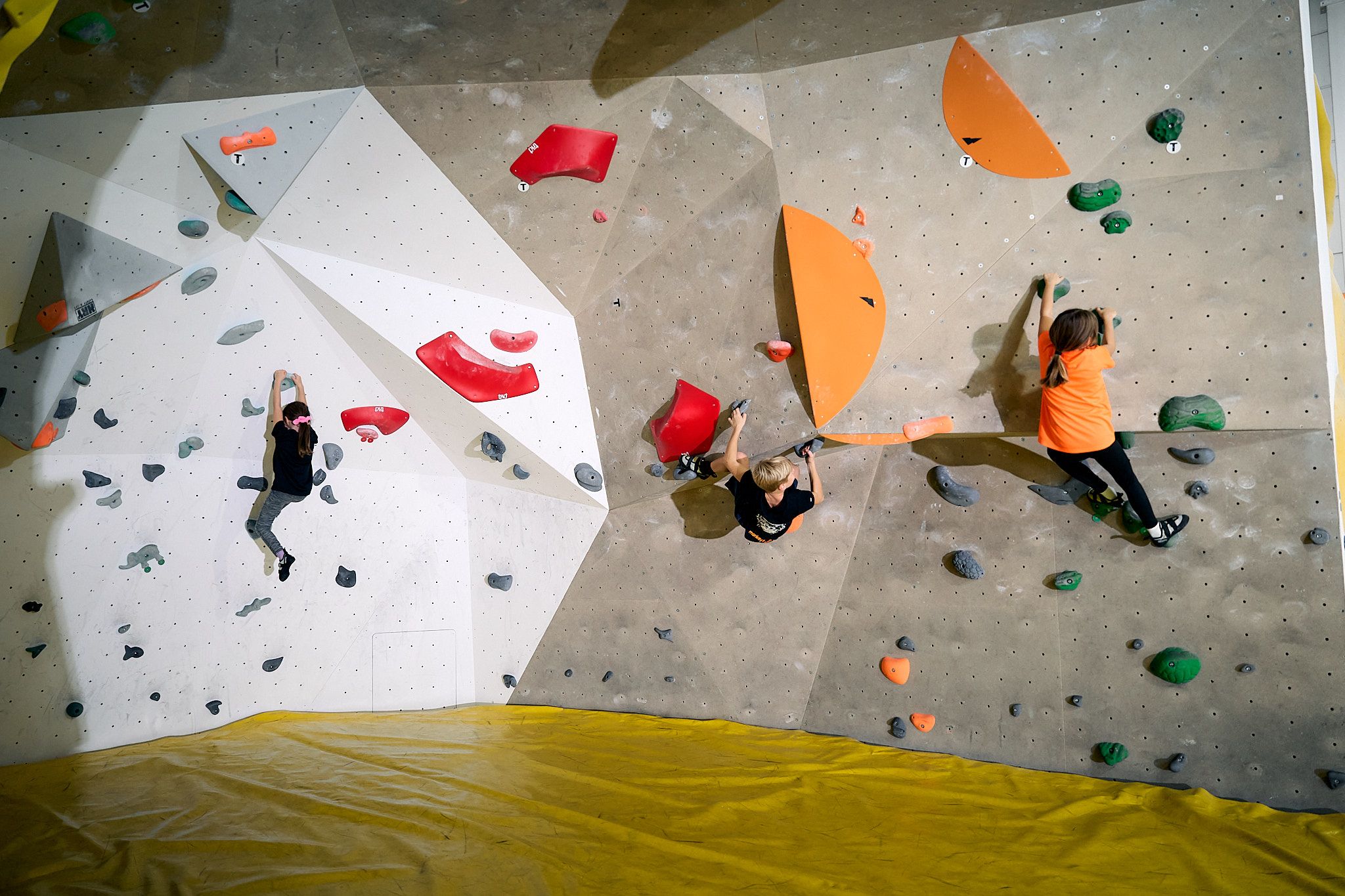 Three children climbing on an indoor bouldering wall with colorful holds.